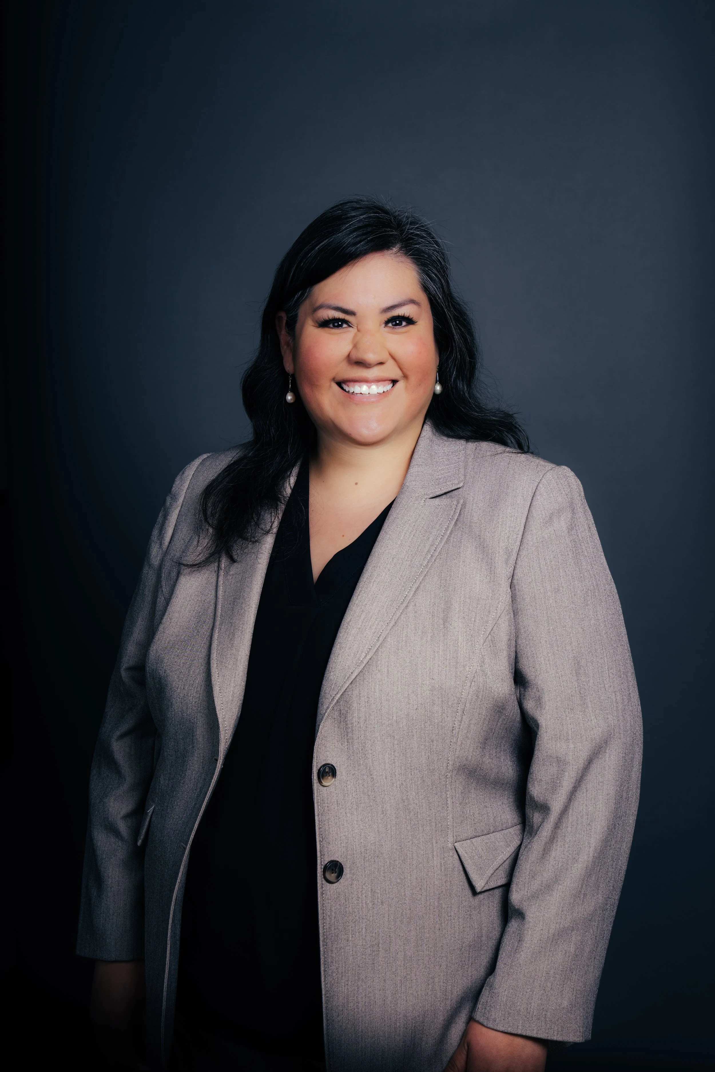 Portrait of a woman with dark hair, smiling, wearing a beige blazer and pearl earrings, against a dark background.