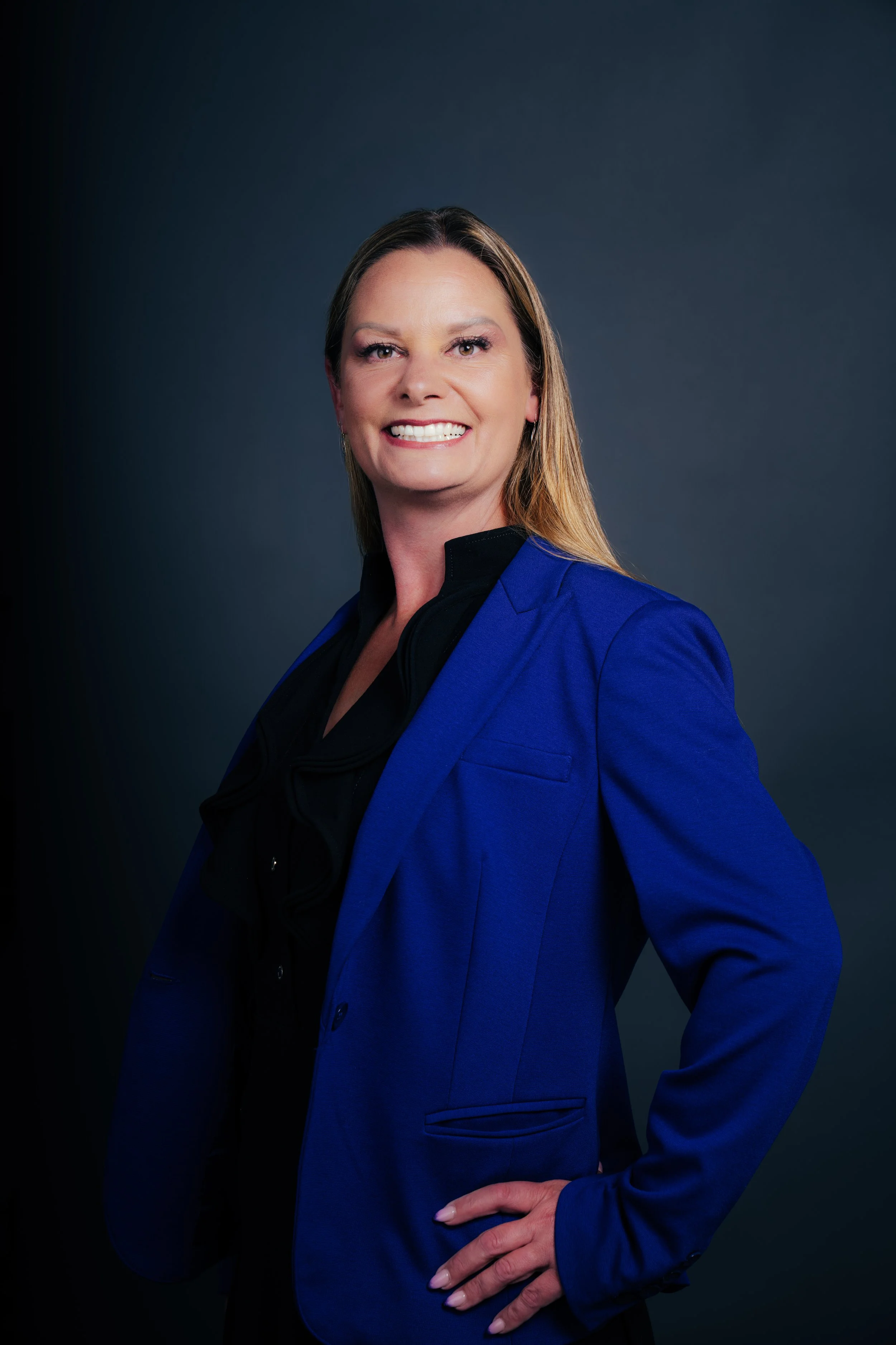 A professional woman with long blonde hair, wearing a bright blue blazer over a black top, poses against a dark gray background with her hand on her hip, smiling confidently.