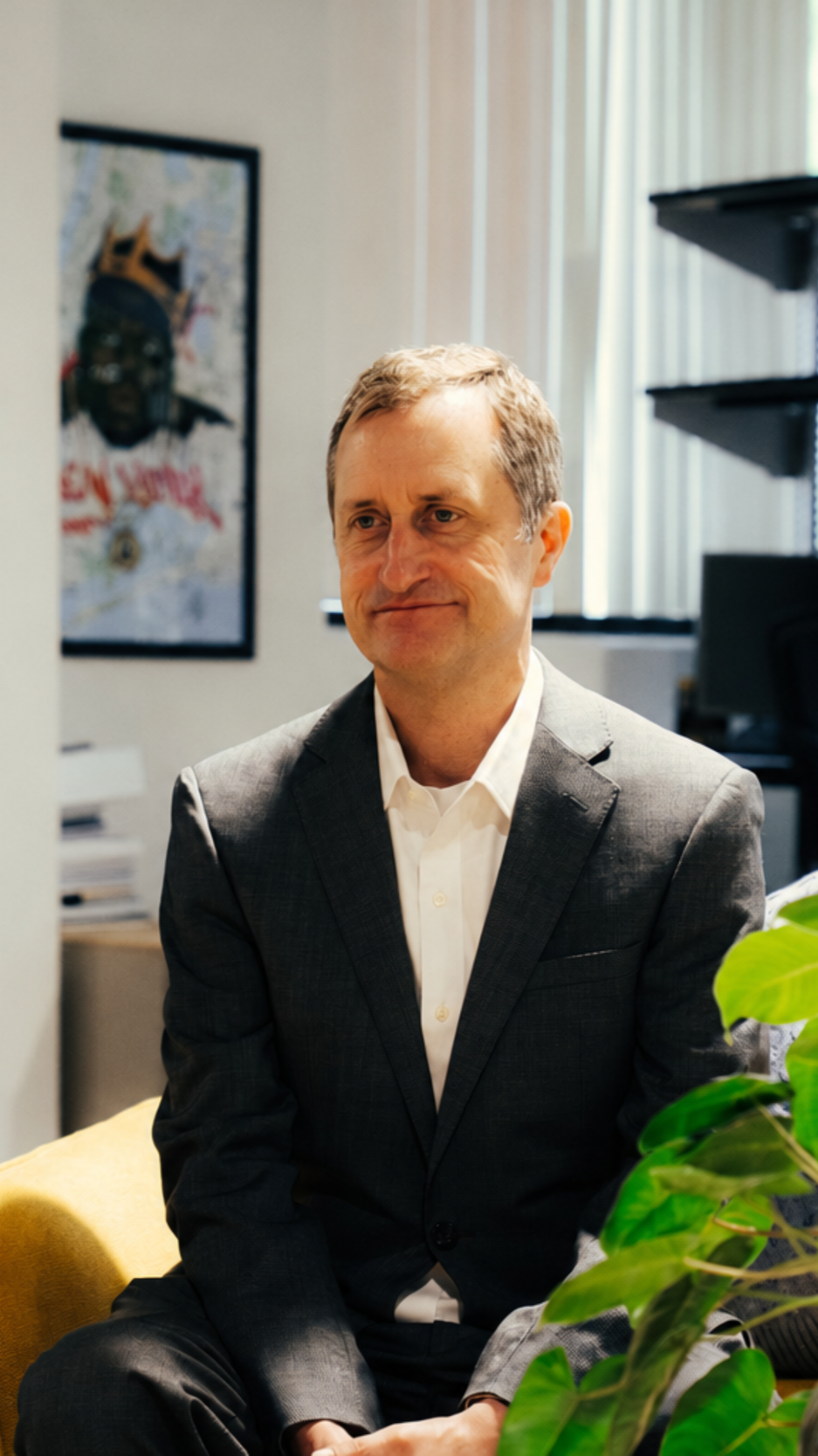 A middle-aged man in a gray suit and white shirt sitting in an office, with a colorful map and a portrait on the wall behind him, and a plant partially visible in the foreground.