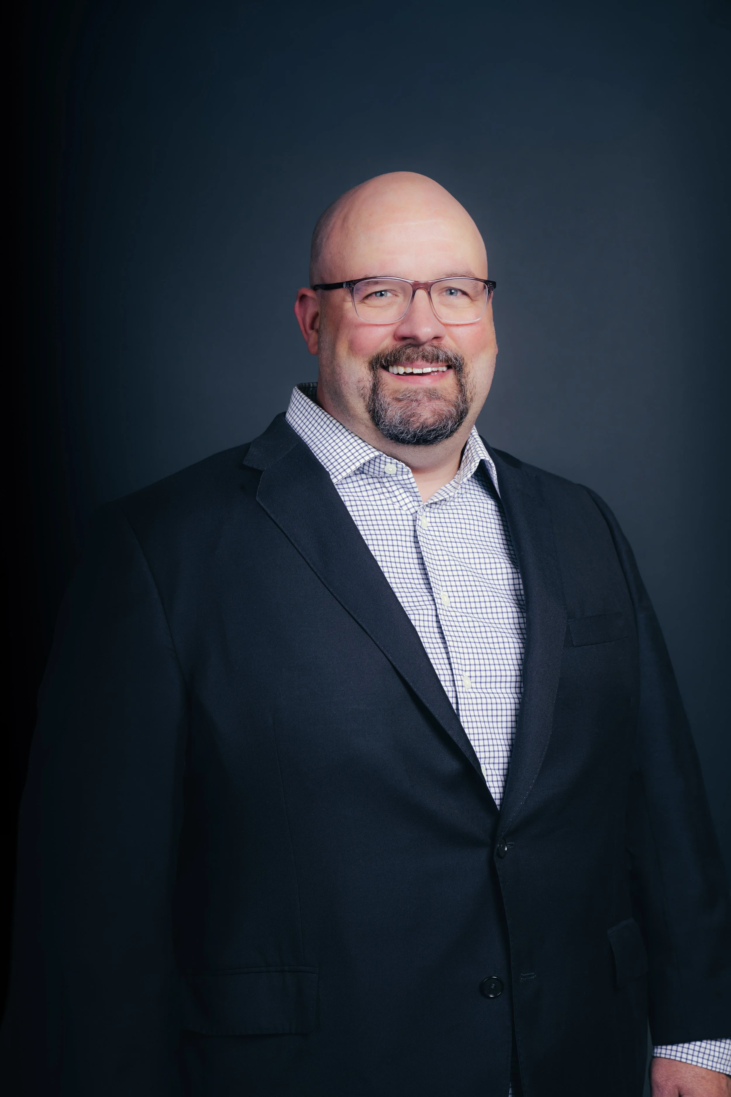 Headshot of a smiling bald man with glasses wearing a black suit and checkered shirt, against a dark grey background.