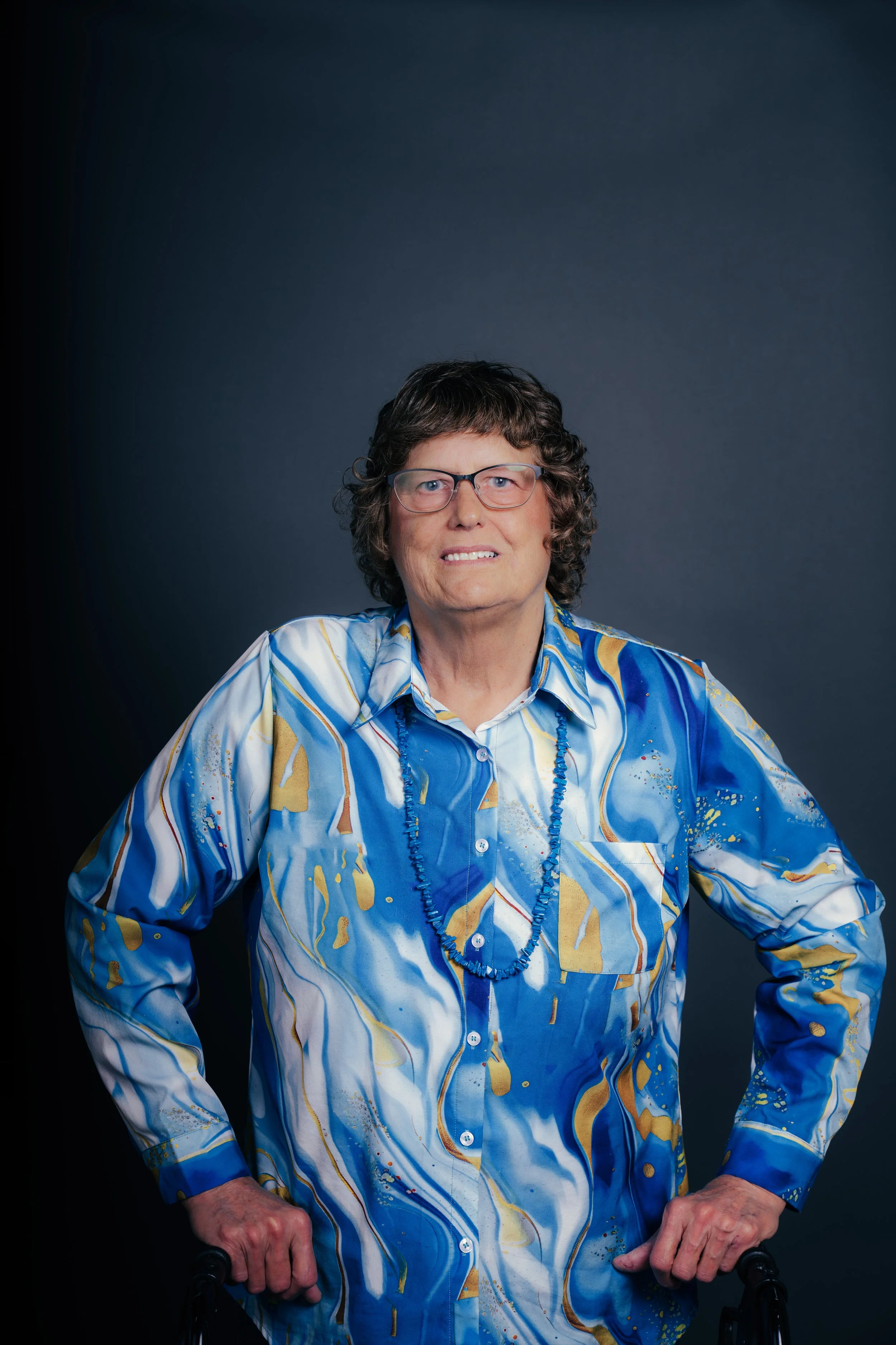 An elderly woman with short, curly hair and glasses, wearing a colorful blue, yellow, and white patterned blouse and a matching necklace, standing against a dark gray background.