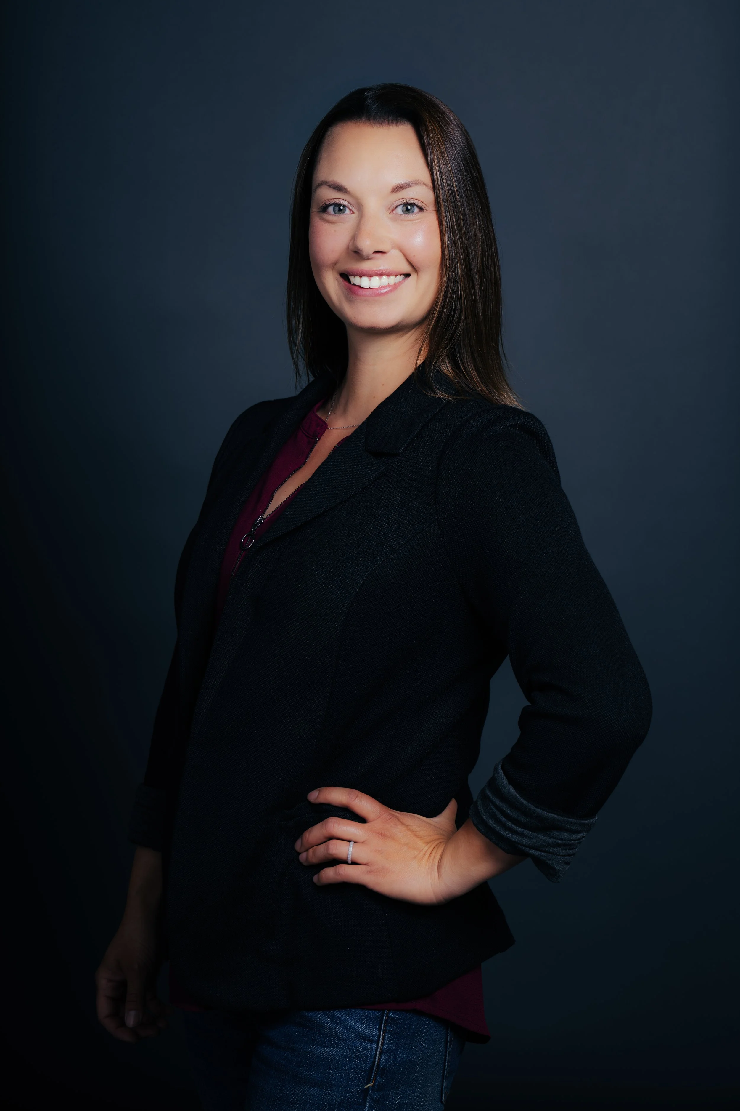 Portrait of a smiling woman with shoulder-length dark hair, wearing a black blazer and blue jeans, standing against a dark background.
