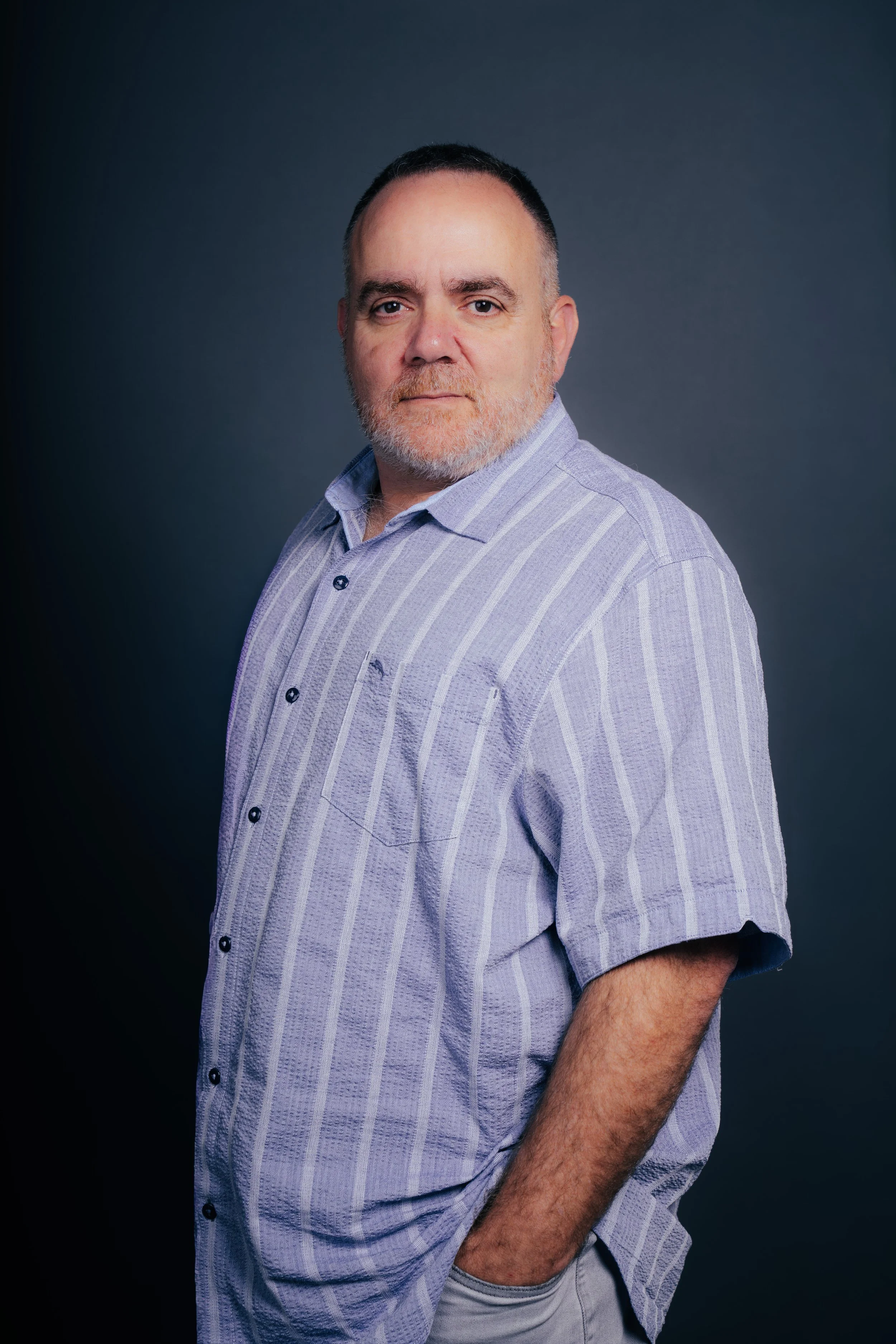 A middle-aged man with a beard and short hair wearing a light purple striped short-sleeve shirt, standing against a dark gray background.