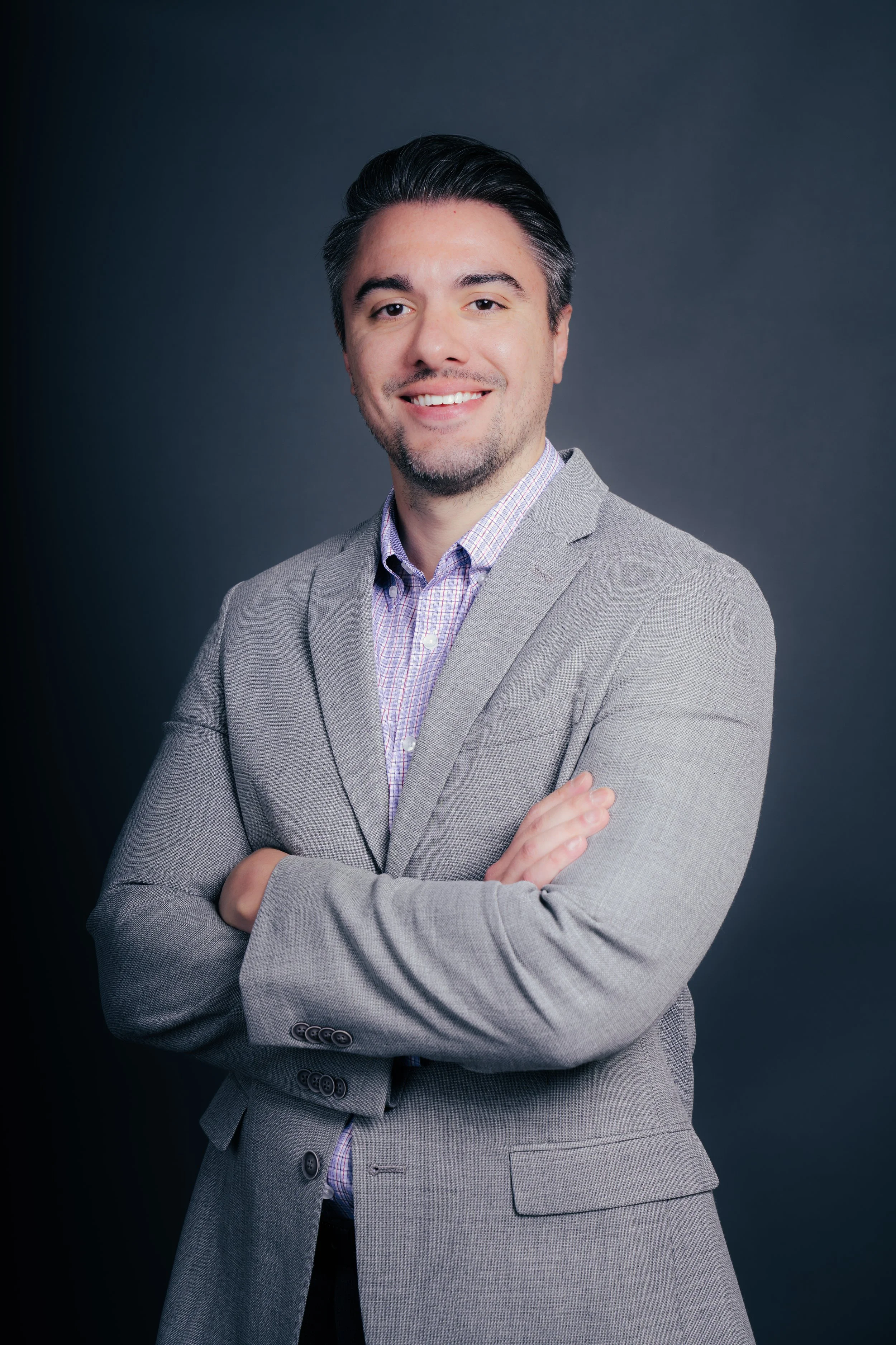 A man with dark hair, wearing a light gray suit with a checked shirt, smiling and crossing his arms, against a dark gray background.