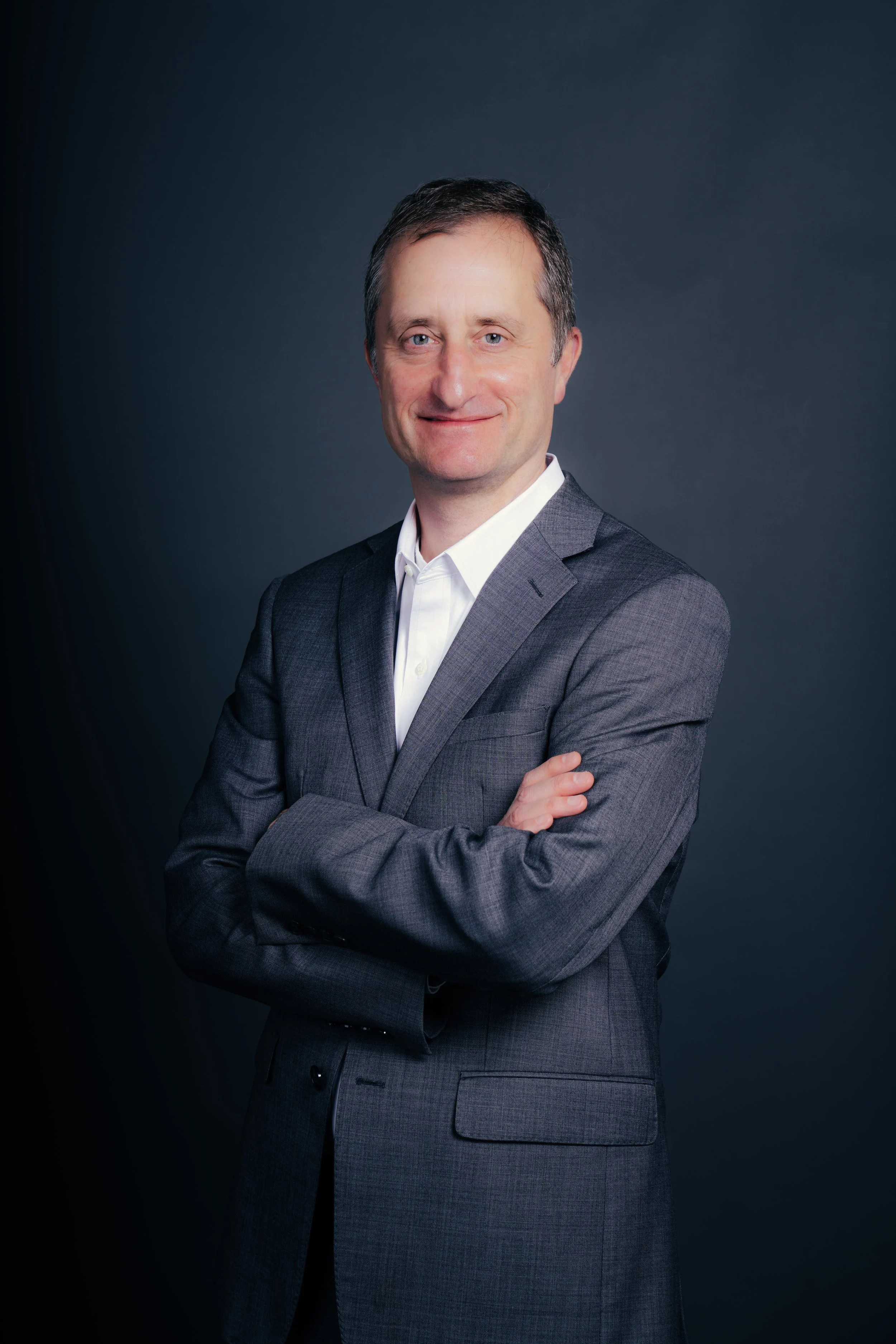 A professional man in a gray business suit and white shirt standing against a dark gray background with his arms crossed, smiling at the camera.