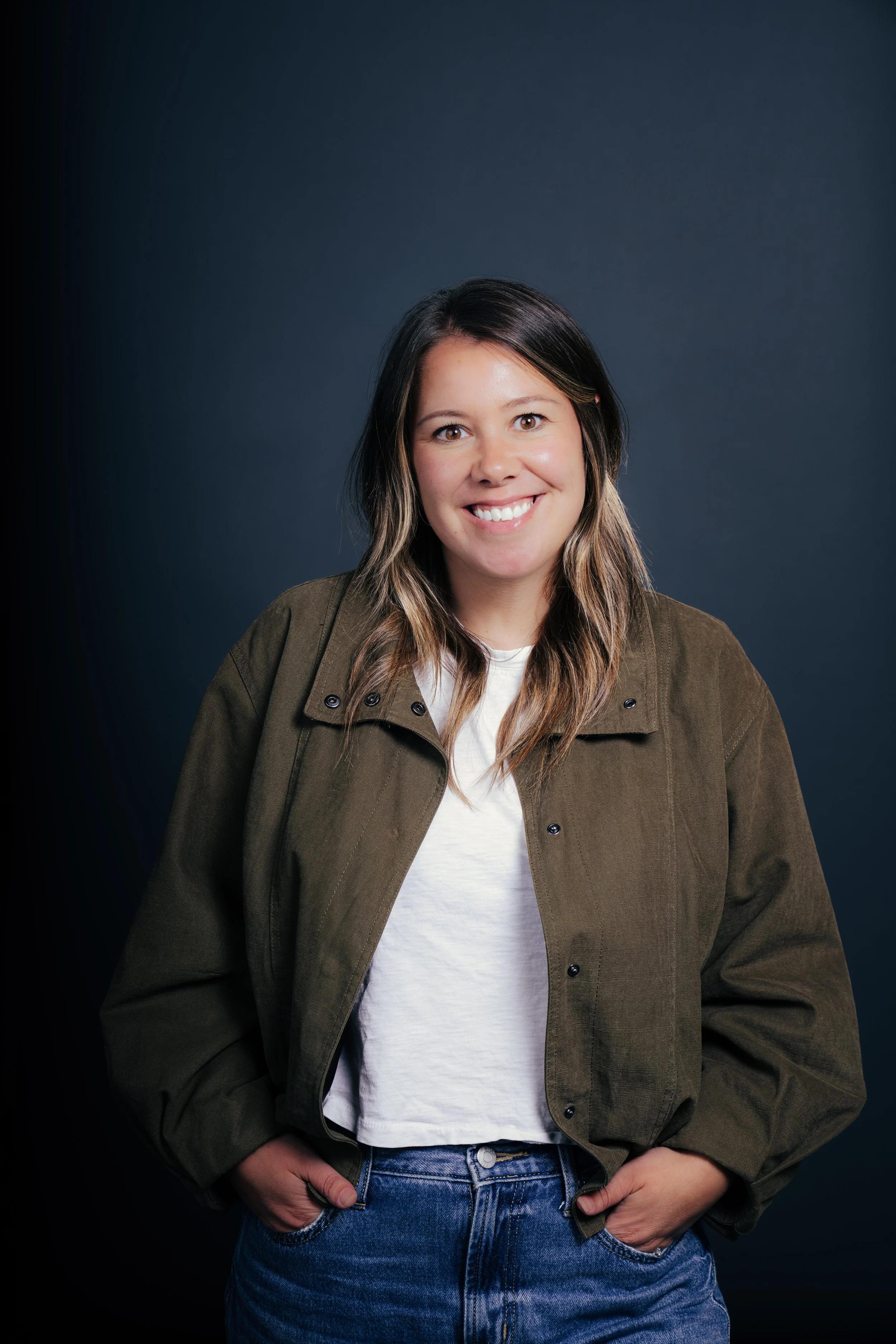 A woman with long brown hair, wearing a brown jacket over a white t-shirt and blue jeans, smiling at the camera with hands in her pockets against a dark background.
