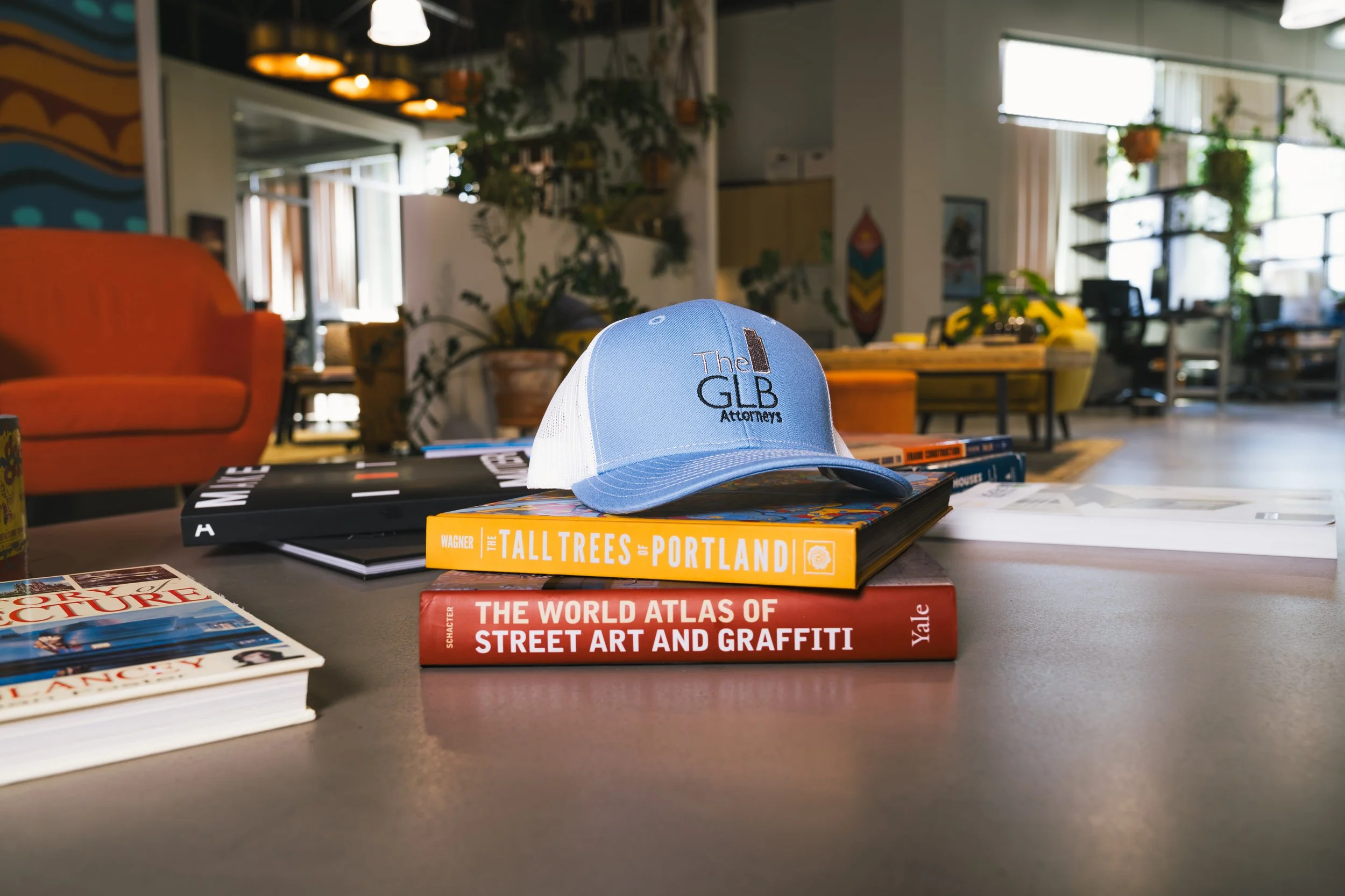 A blue and white baseball cap with the text 'The GLB Attorneys' placed on top of a stack of books on a table in a cozy indoor setting.