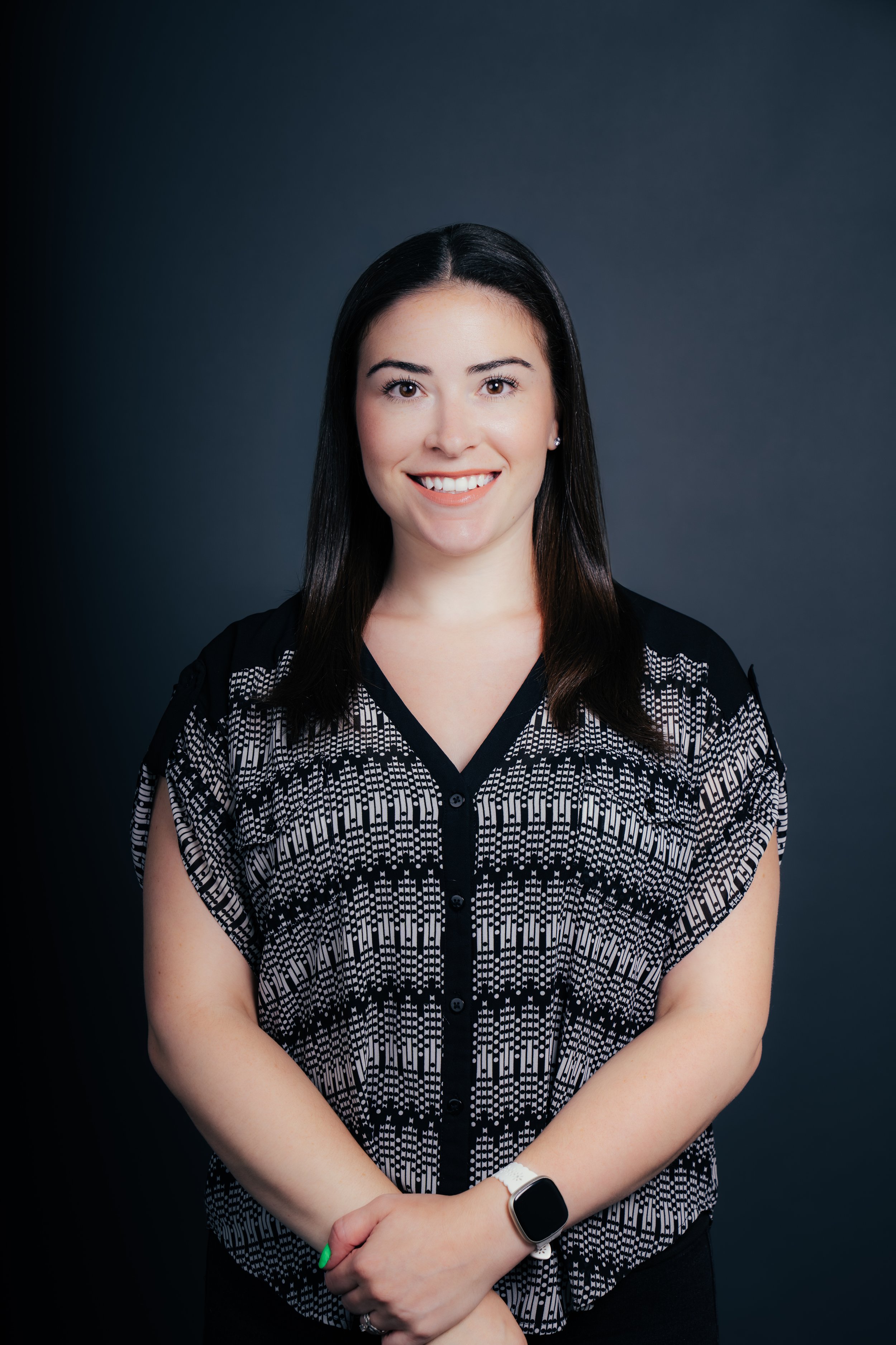 A woman with dark hair smiling, wearing a black and white patterned blouse, standing against a dark background.