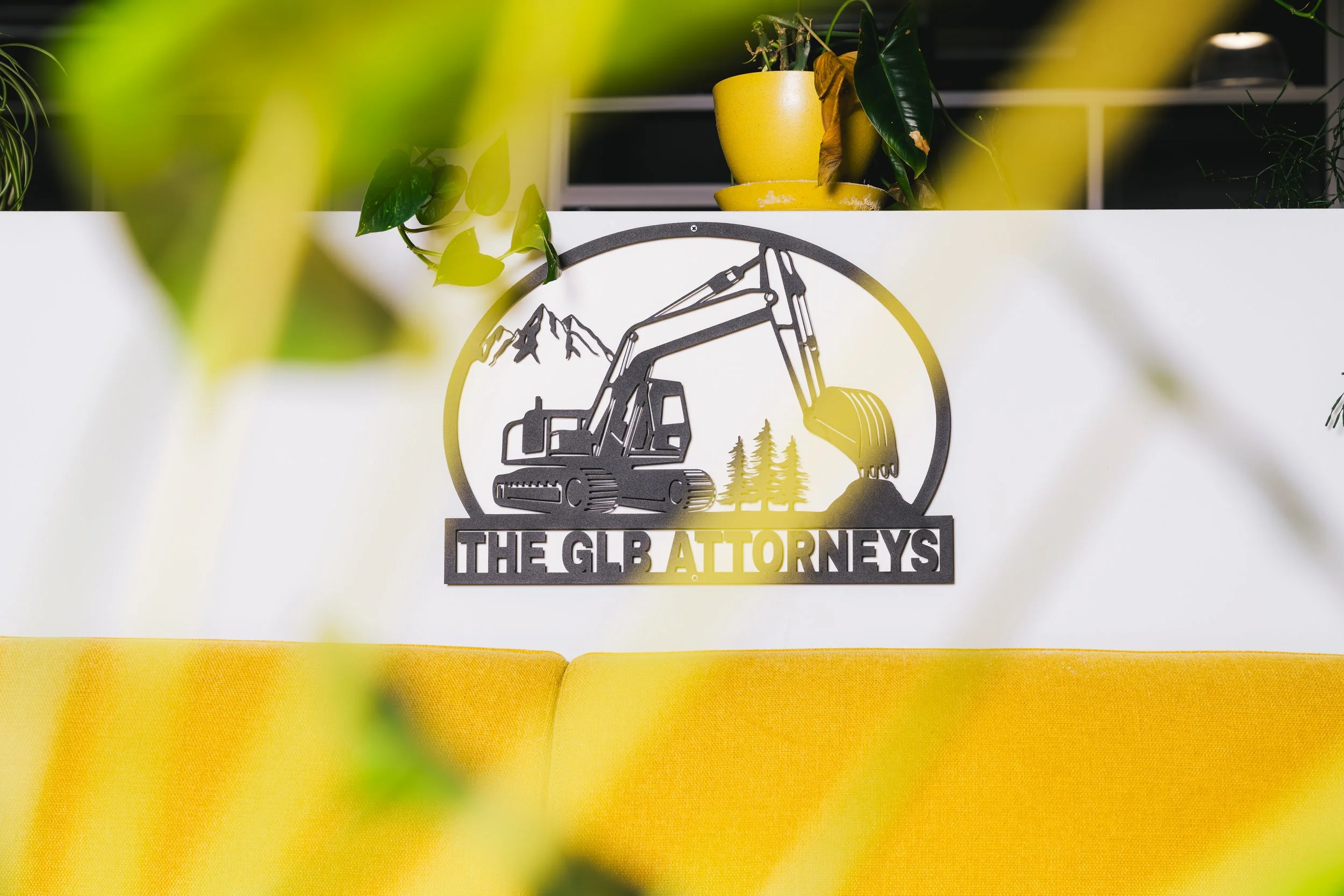A sign with a mountain and excavator logo that reads "The GLB Attorneys" is on a white wall. There are yellow plant pots with green plants above the logo. Yellow fabric covers the furniture below the sign, and some yellow leaves are blurred in the foreground.