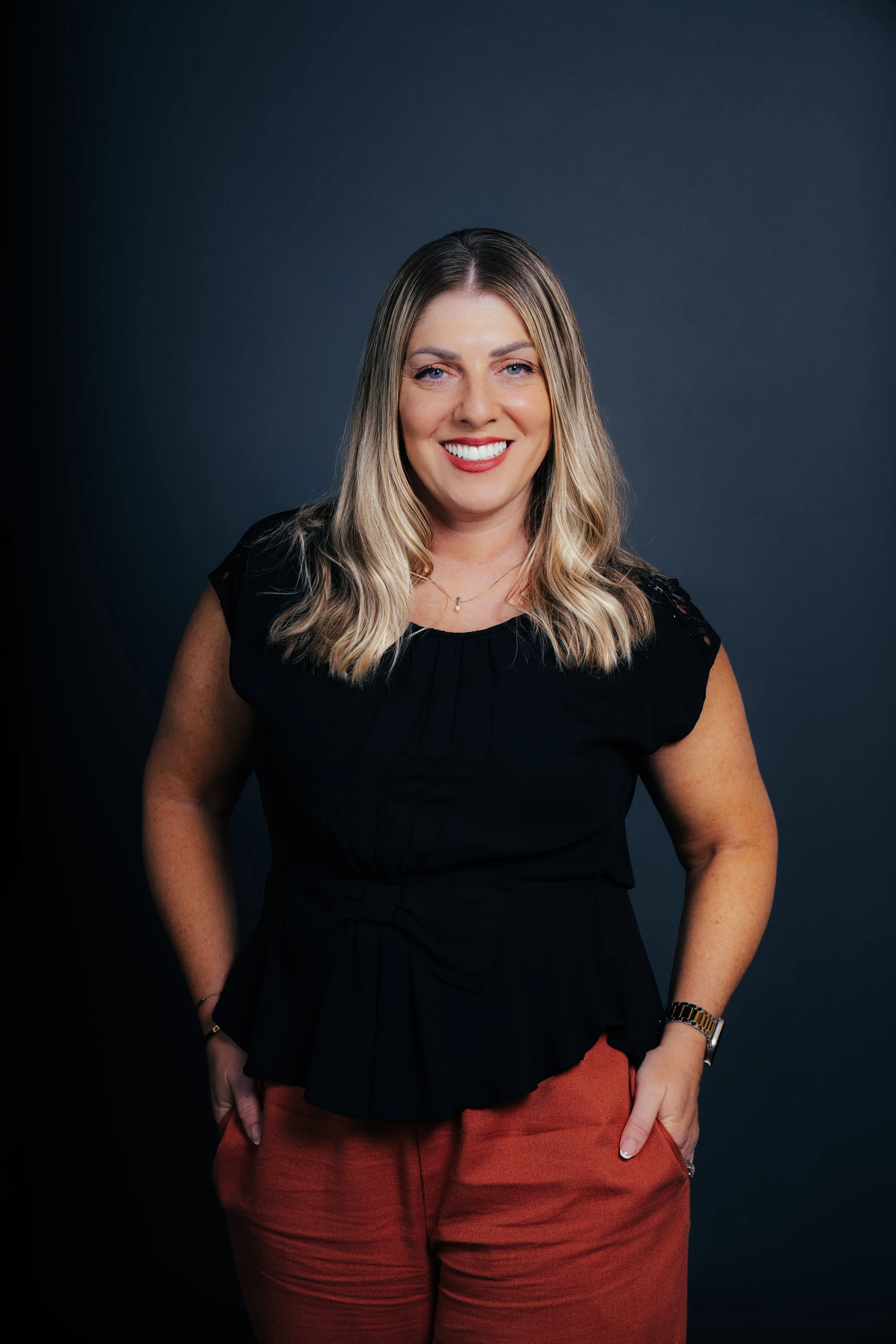 A woman with blonde hair and blue eyes smiling, wearing a black top, orange pants, a watch, and a necklace, standing against a dark gray background.
