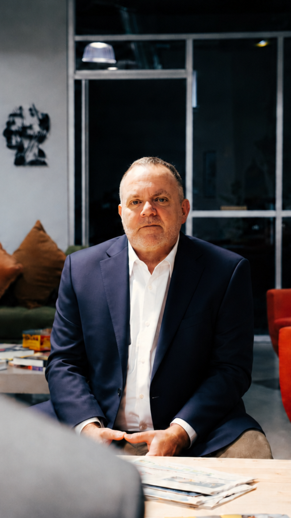 A middle-aged man in a dark suit and white shirt sitting at a table in a modern office or meeting room, with a serious expression. There are cushions on a sofa and a bookshelf with magazines in the background.