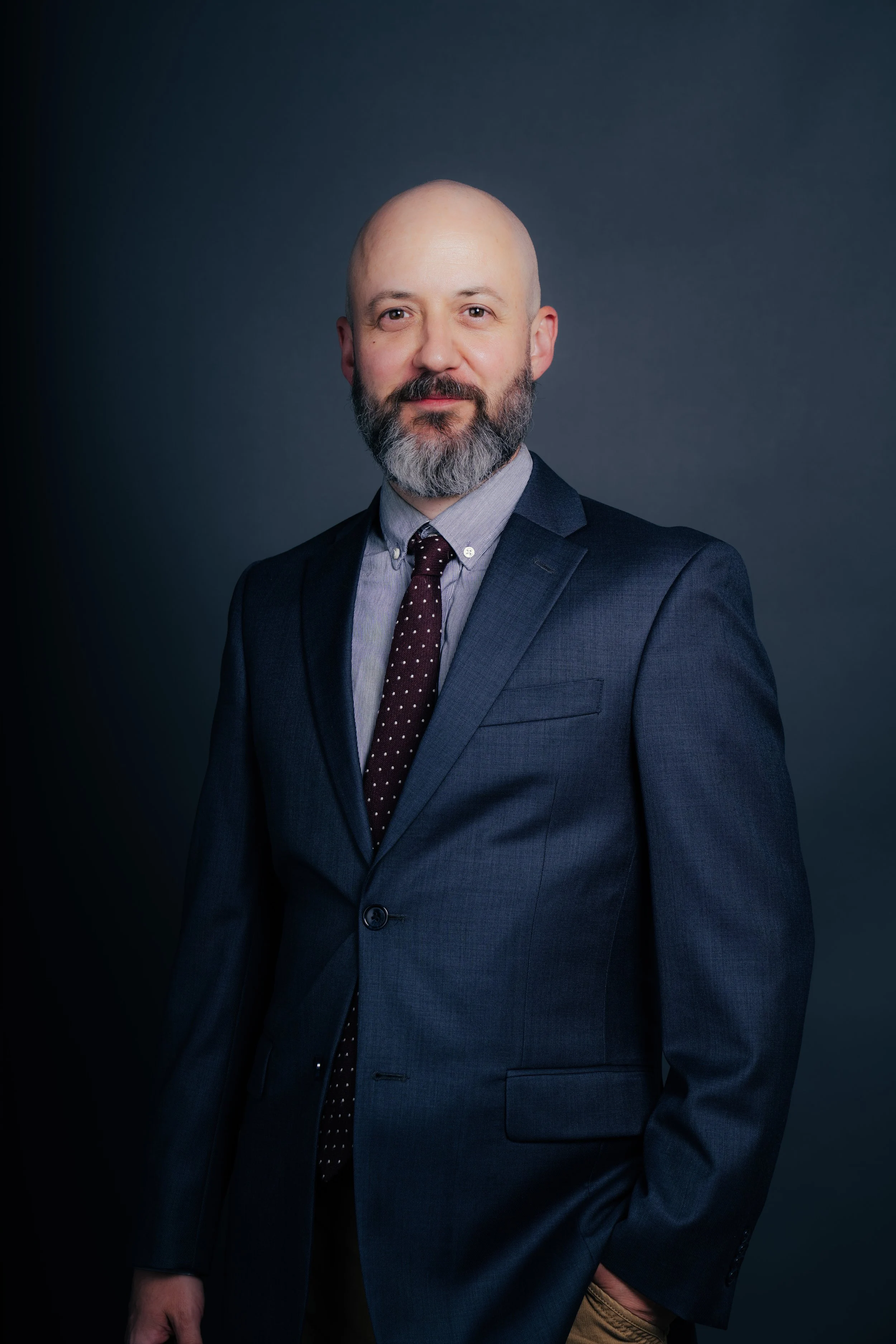 A well-dressed man with a beard and bald head, wearing a dark suit, light shirt, and patterned tie, standing against a dark background.