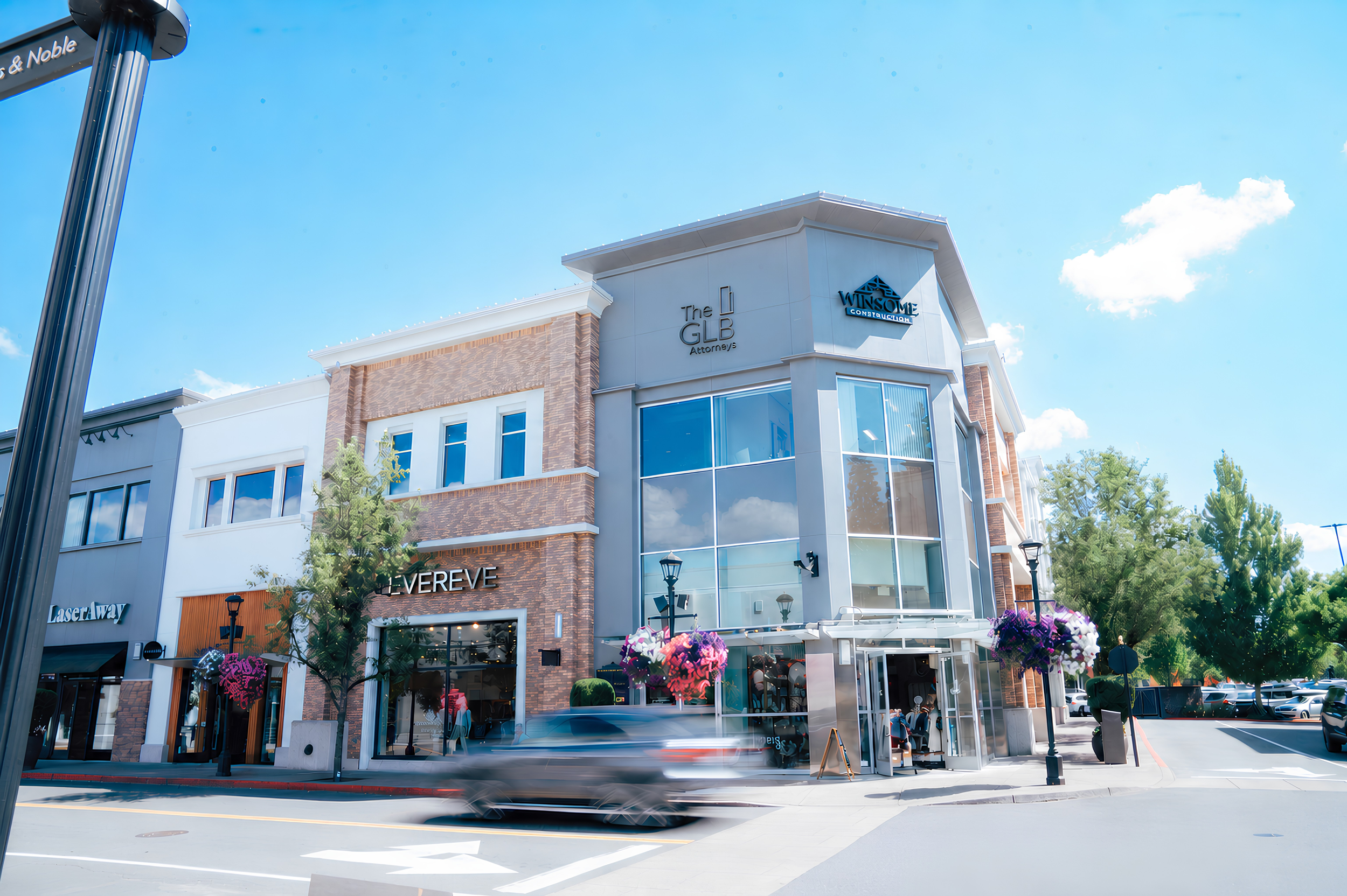 Modern commercial building with retail shops on the ground floor, including a store named 'LE VERVE', and signs for 'The GLB Attorneys' and 'WINSOME CONSTRUCTION'. There are flowering plants, trees, street lamps, and cars parked outside on a sunny day with blue skies and some clouds.