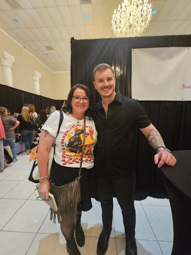 A smiling woman with glasses and a man in black shirt posing together at an indoor event with a large chandelier and black curtains in the background.