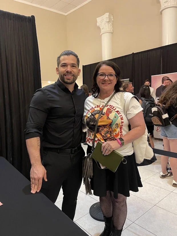 A man and a woman standing together indoors, smiling at the camera. The man has dark hair, a beard, and is wearing a black shirt and black pants. The woman has glasses, dark hair, and is wearing a white graphic t-shirt, a black skirt, and fishnet stockings. She is holding a green notebook and is wearing a pink wristband.