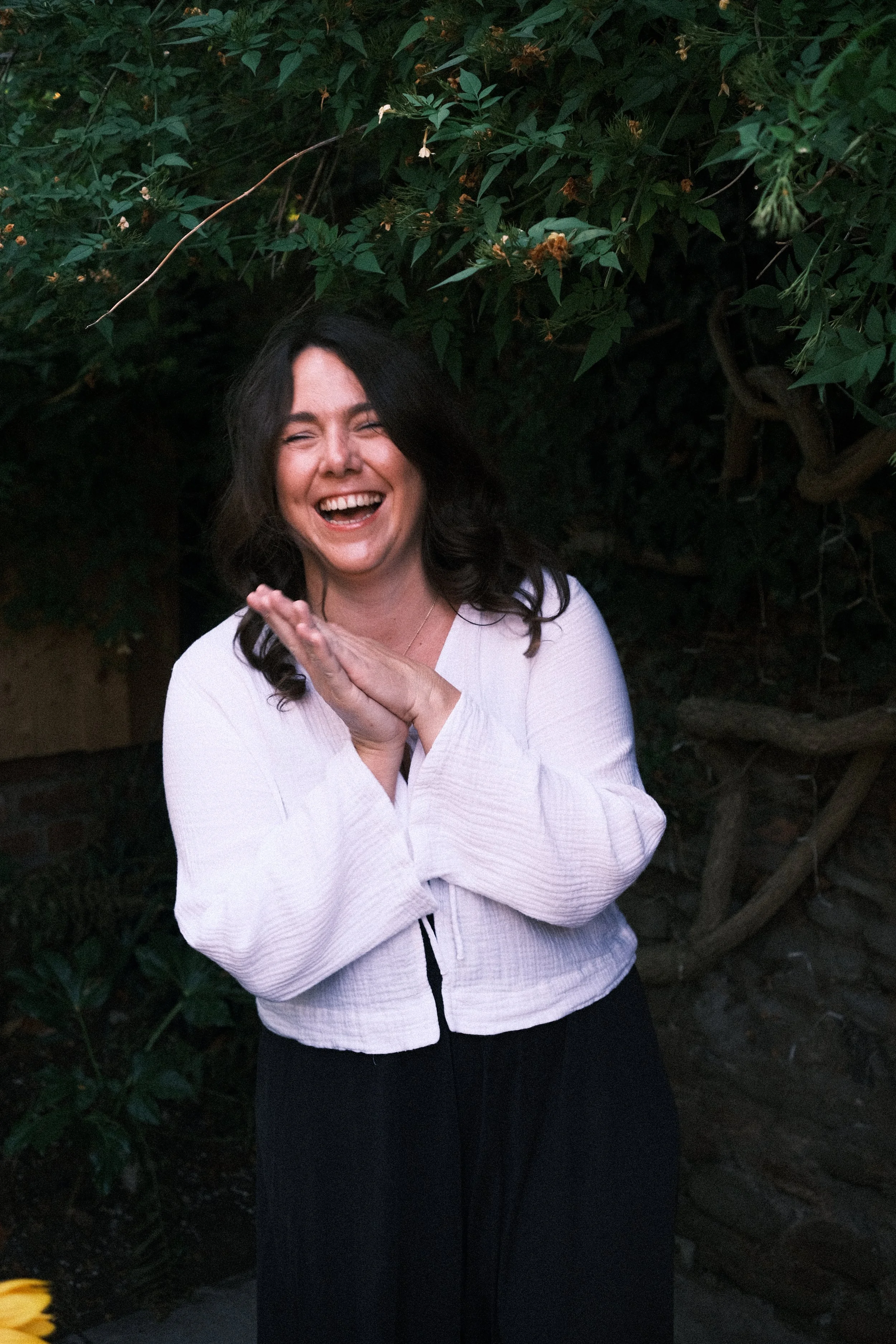 A woman with dark hair and a white jacket laughing outdoors, standing in front of a leafy green hedge.