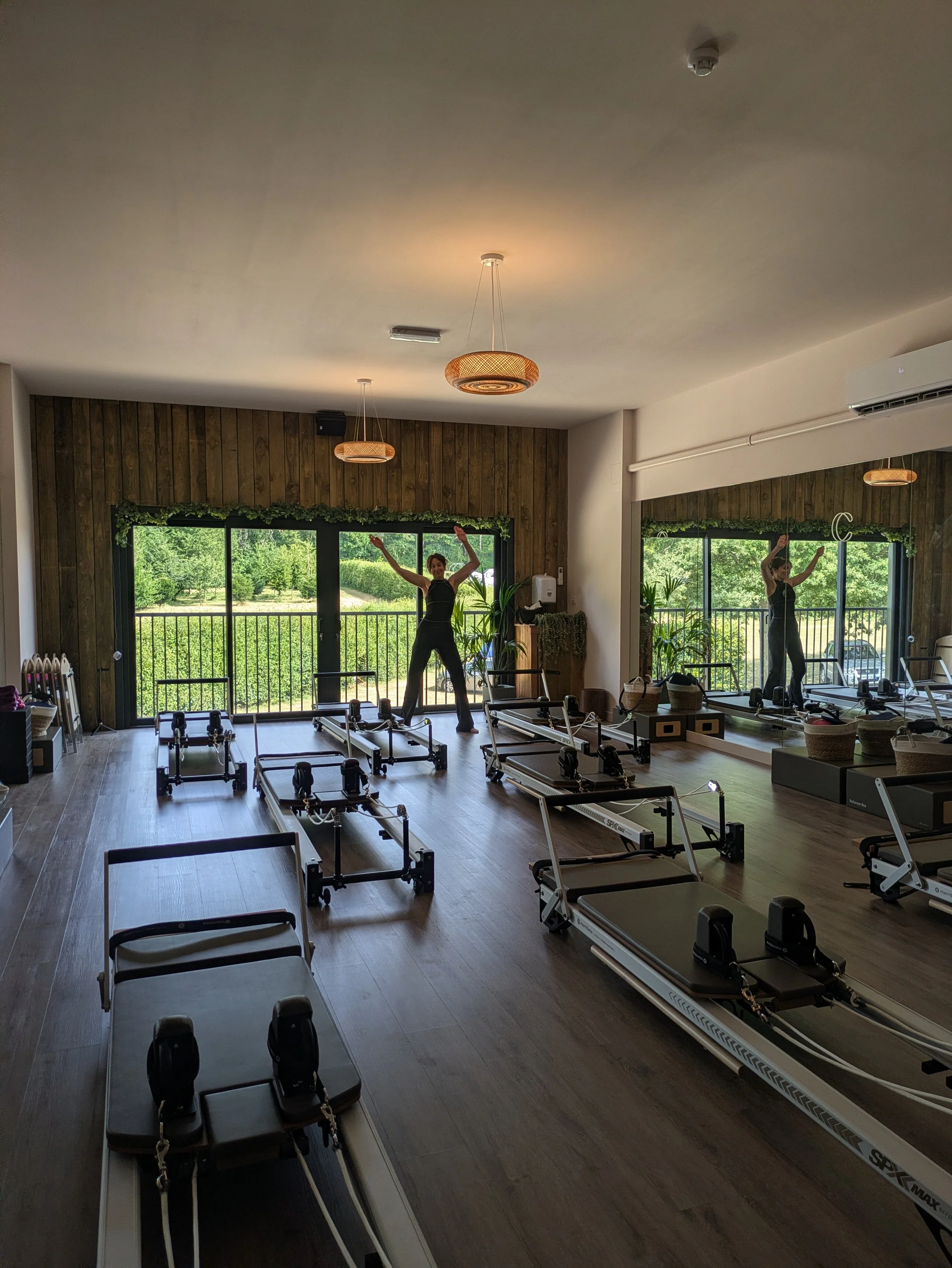 Interior of a reformer Pilates studio with Pilates reformer machines, two women exercising, large windows showing a green outdoor landscape, and ceiling lights.