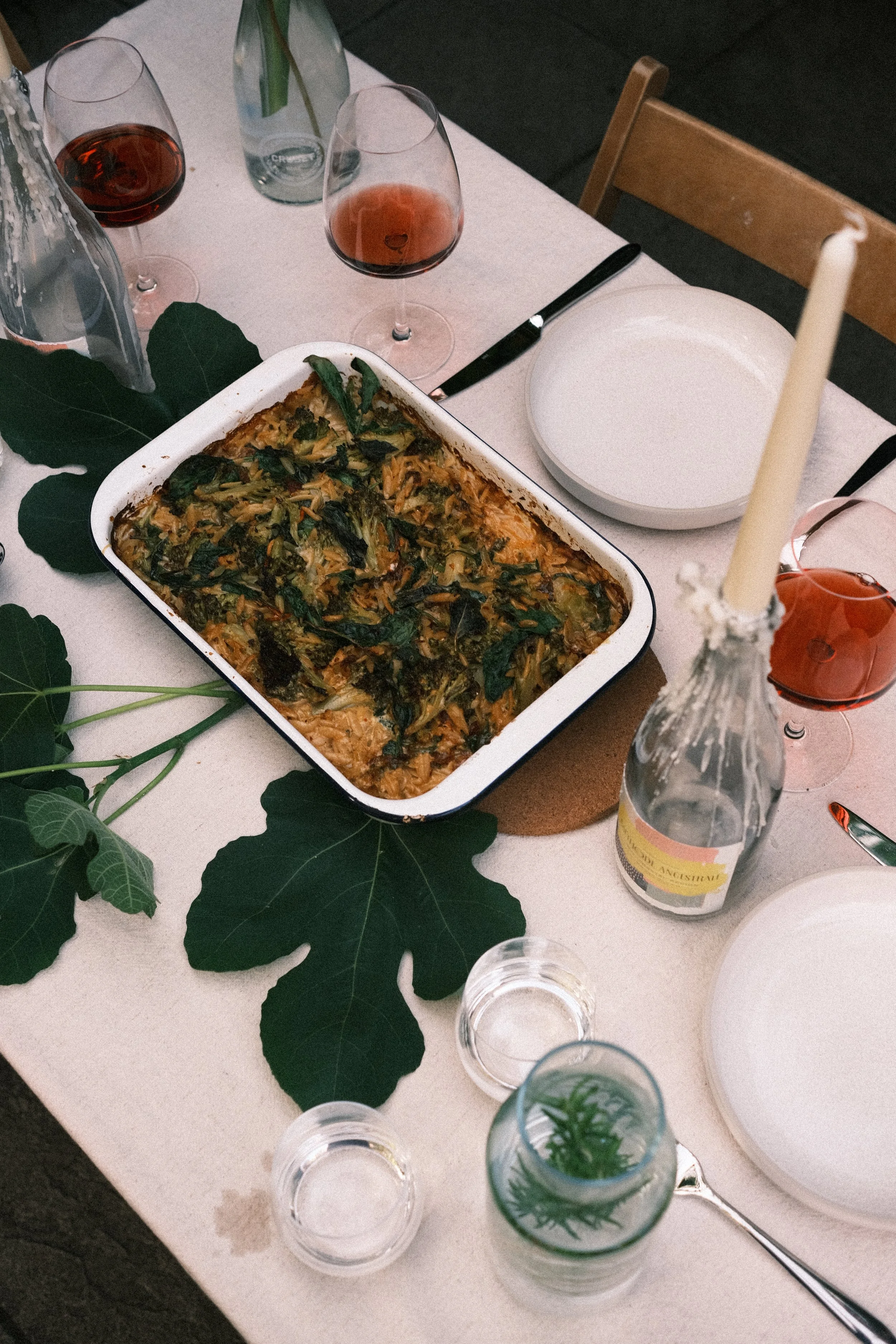 Table set with a casserole dish of leafy vegetable casserole, two glasses of red wine, various water bottles, a charger plate, and a candle, with green leaves decorating the table.