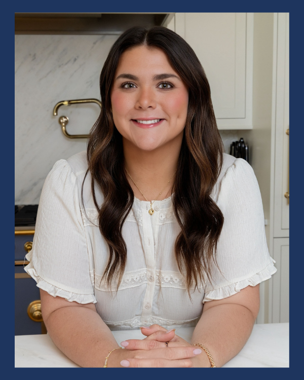 A woman with long brown hair, wearing a white blouse, smiling and sitting at a kitchen counter.