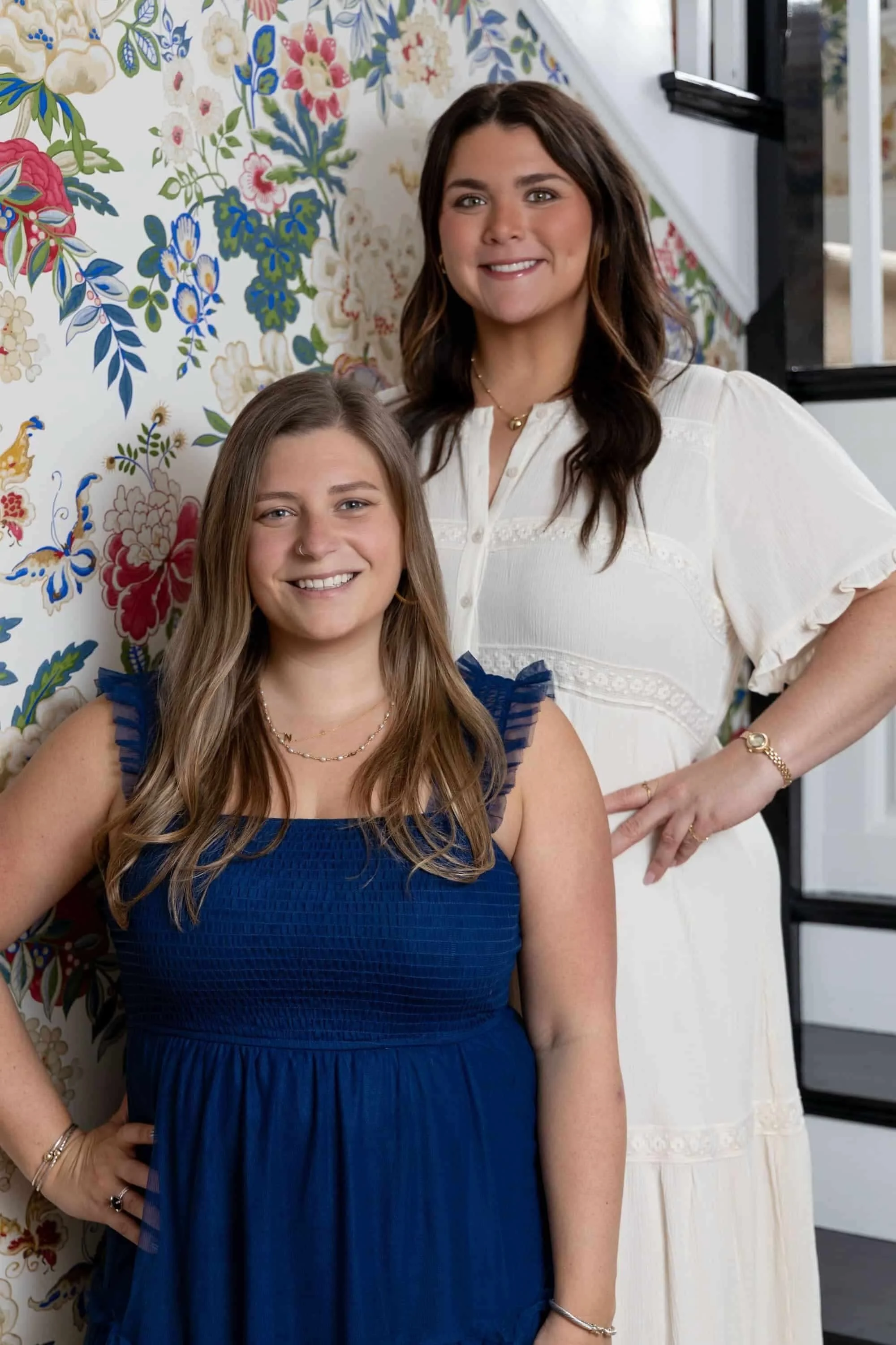 Two women smiling, one wearing a blue dress and the other in a white dress, standing in front of a floral-patterned wall.