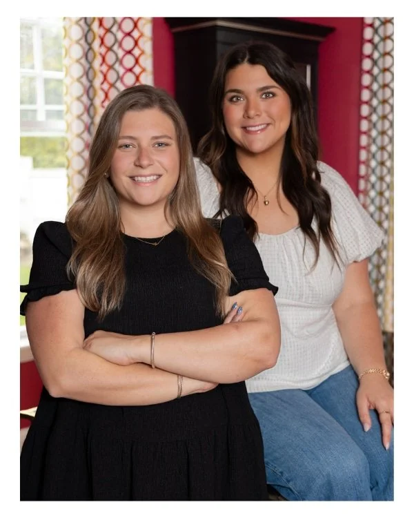 Two women smiling and posing indoors, one with crossed arms in a black dress, the other sitting in a white blouse and blue jeans, with colorful curtains and a window in the background.