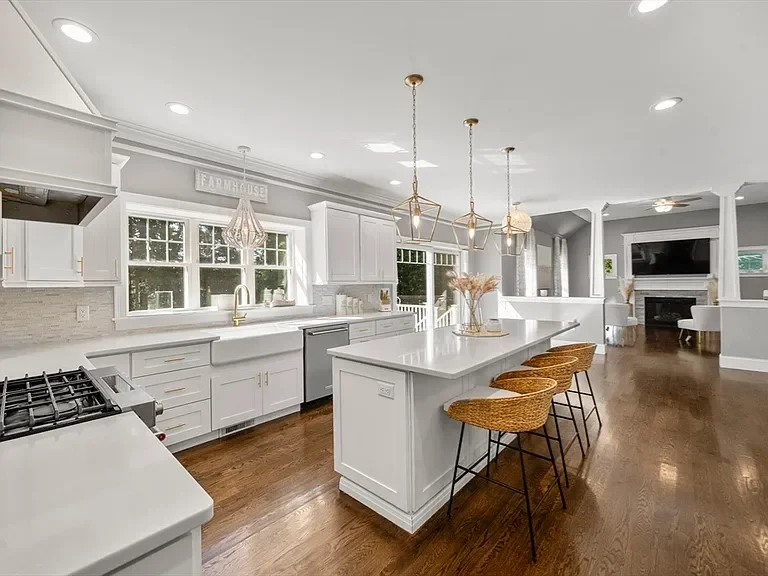 Modern white kitchen with island, wicker bar stools, stainless steel appliances, pendant lights, large windows, and open layout leading to living room with fireplace.