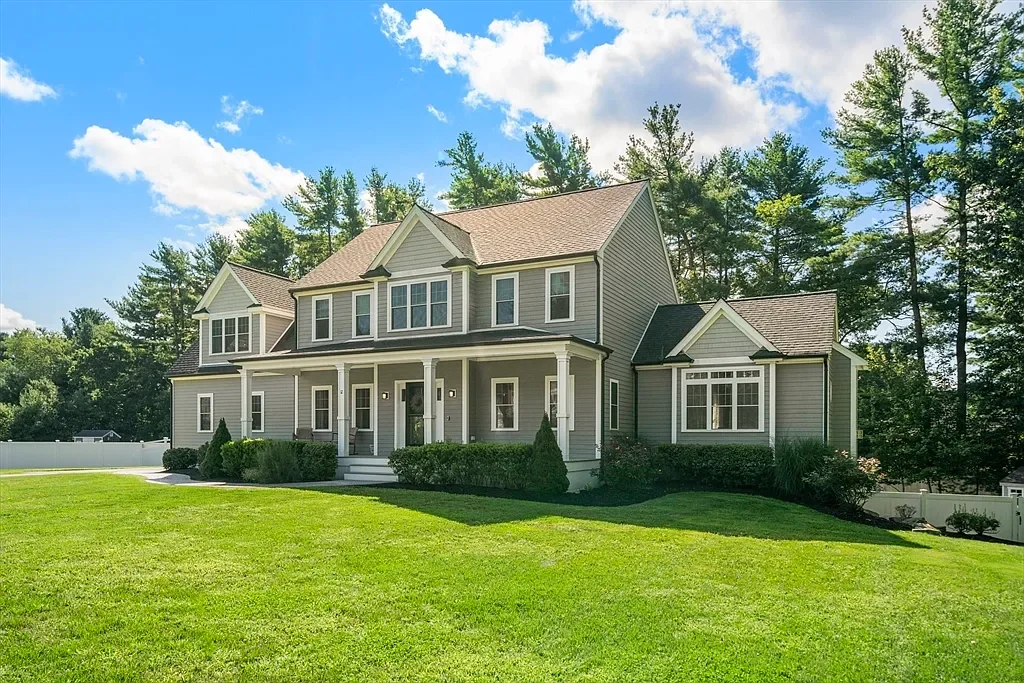 A large two-story house with gray siding, white trim, and a brown roof, surrounded by a green lawn and trees under a partly cloudy sky.