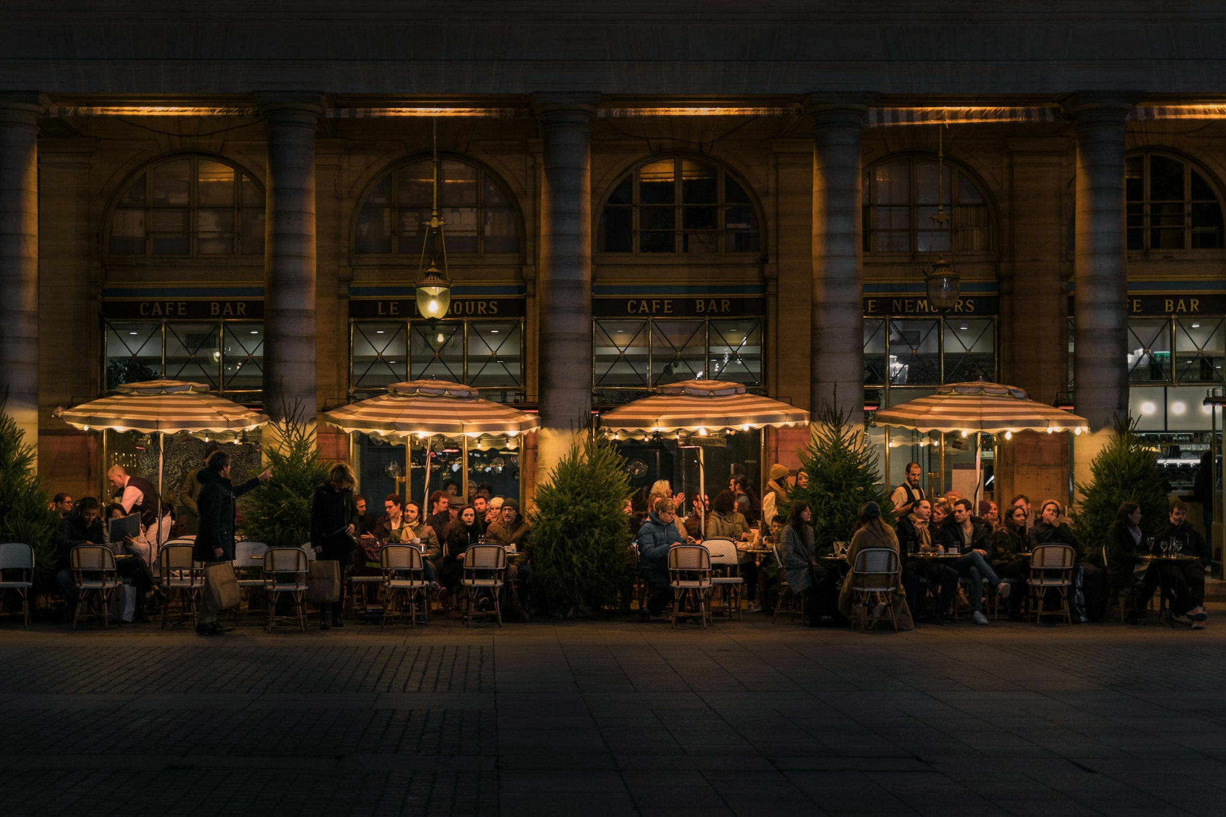 Night scene of an outdoor cafe with people dining and socializing under striped umbrellas in front of a historic building with large arched windows and columns, illuminated by warm street lighting.