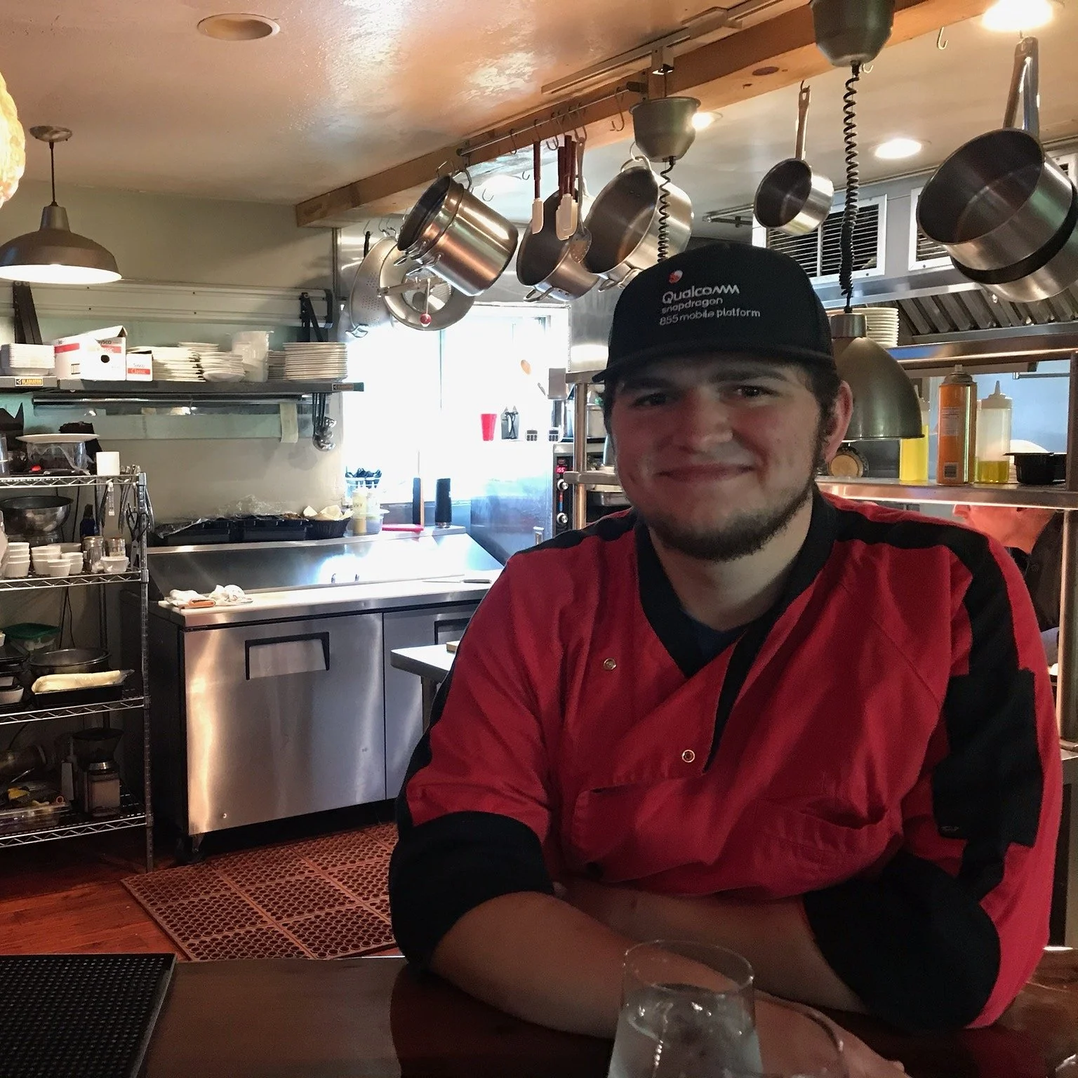 A smiling man wearing a black cap with the Qualcomm Snapdragon logo and a red chef jacket, sitting in a restaurant kitchen with hanging pots and shelves of dishes in the background.