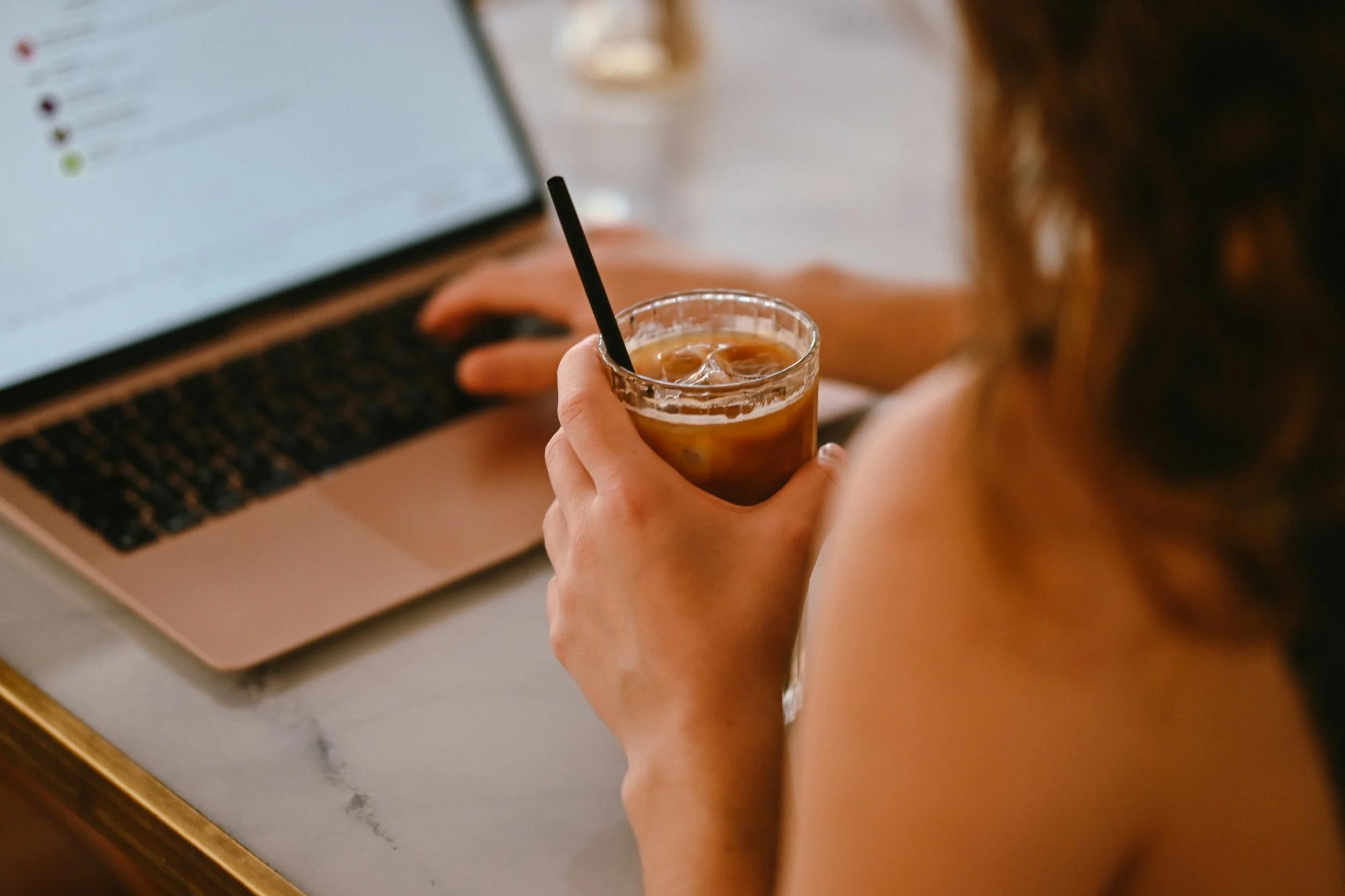 Woman seen from behind the shoulder with one hand on her laptop and the other holding an iced coffee in soft light, representing starting a task at the last minute