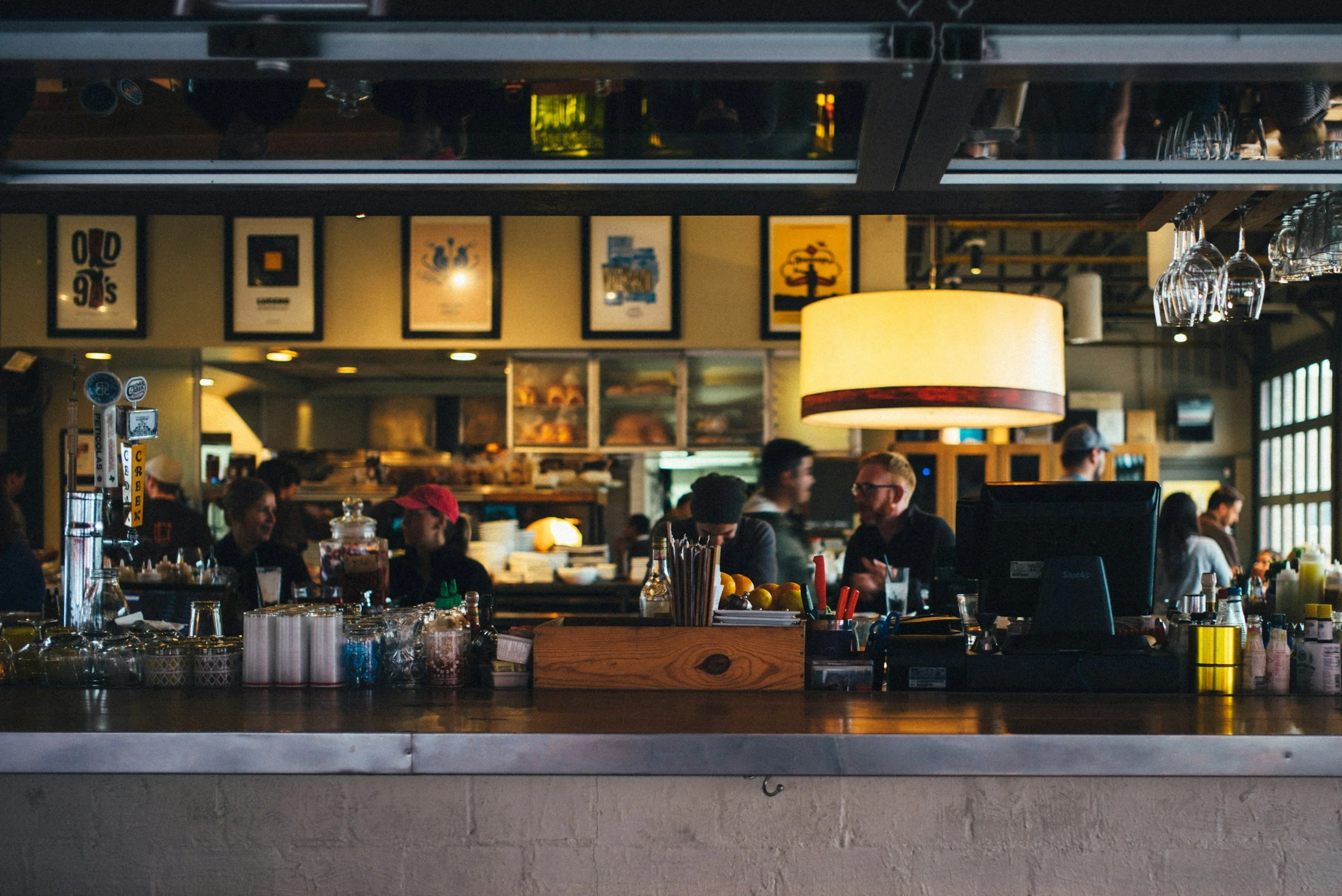 Inside a busy restaurant or bar, with patrons seated at the counter and artwork on the wall, glasses hanging upside down, and beer tap handles visible.