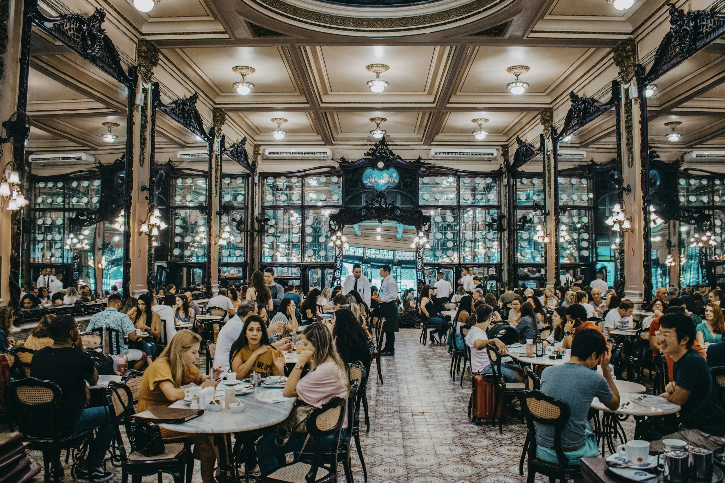 A crowded indoor restaurant with many people seated at tables, waiters serving, ornate mirrors and chandeliers on the ceiling, and a decorative black metal archway with a painted scene at the top.