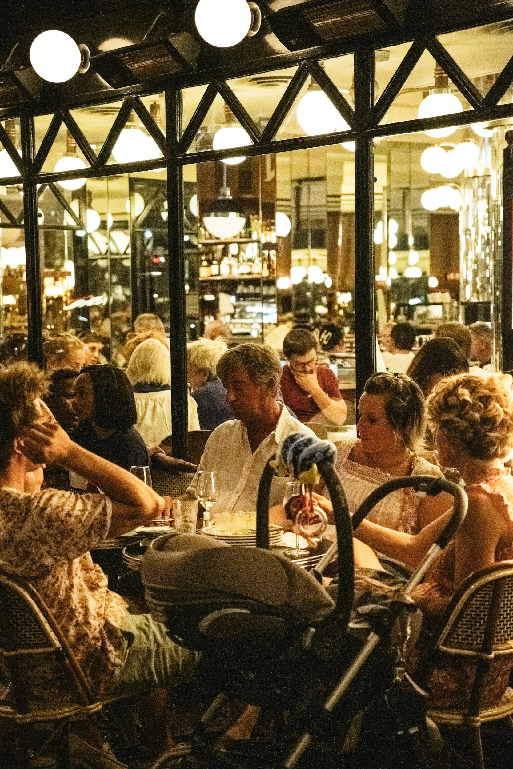 People sitting at tables in a busy, warmly lit restaurant with large glass windows and decorative lighting fixtures.