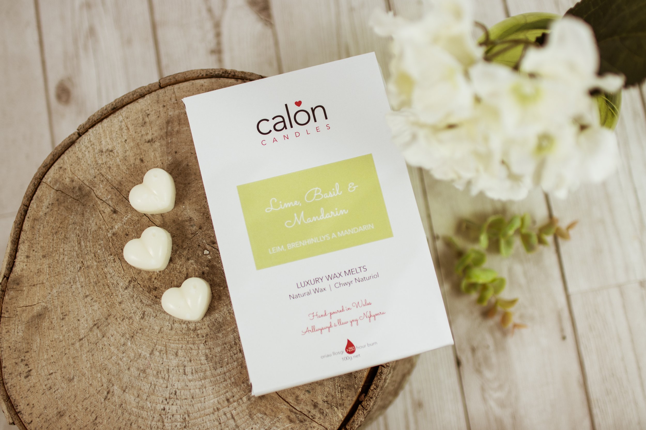 White heart-shaped candles on a wooden surface next to a Calon Candles product packaging, with blurred flowers in the background.