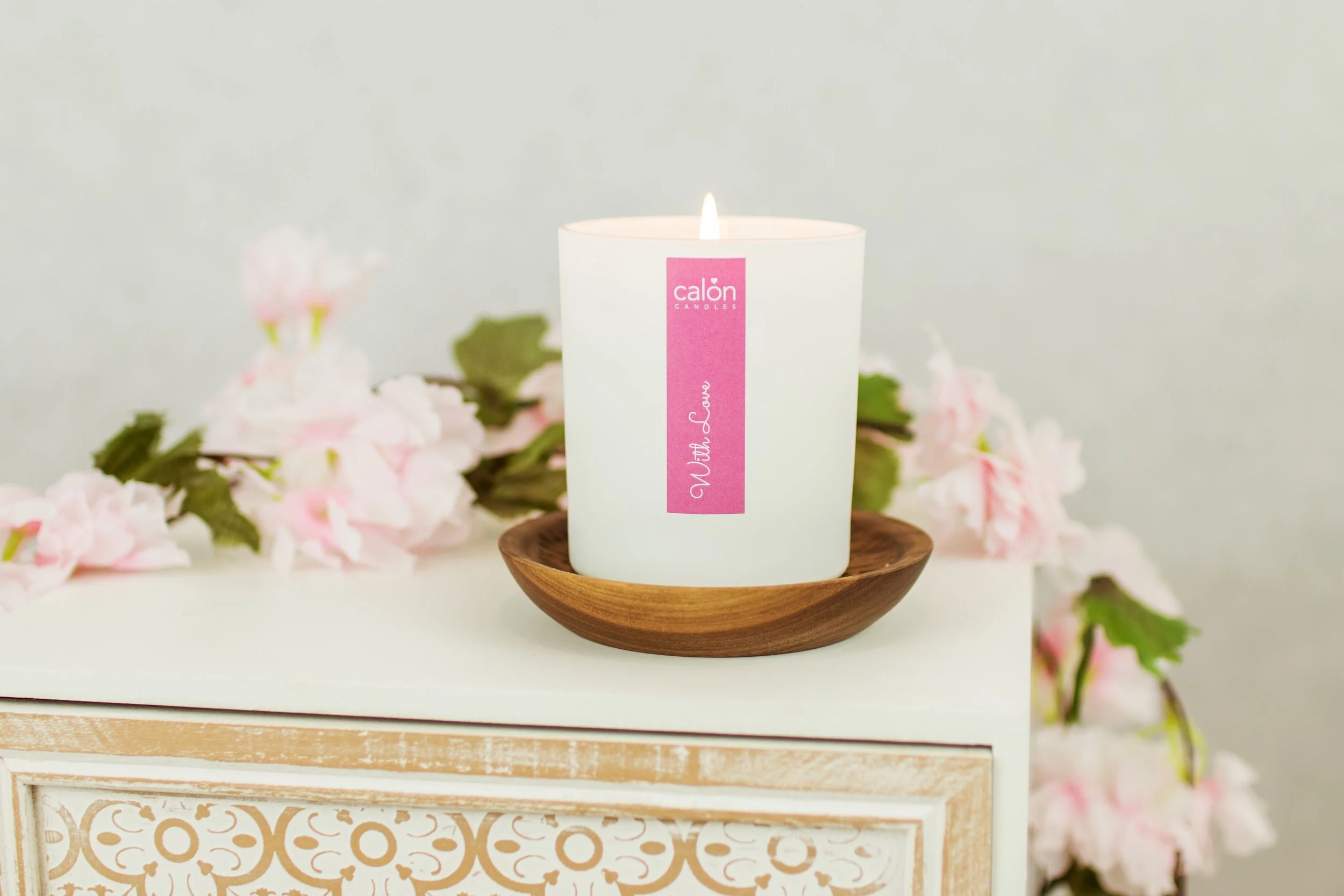 A white candle with a pink label that reads "calon candles" and "with love" is lit and placed in a small wooden bowl. Pink and white flowers are blurred in the background.