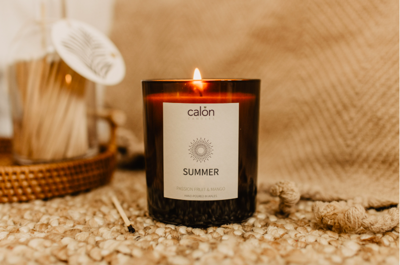 A lit black candle with a white label reading  'SUMMER' on a textured surface with decorative items in the background.