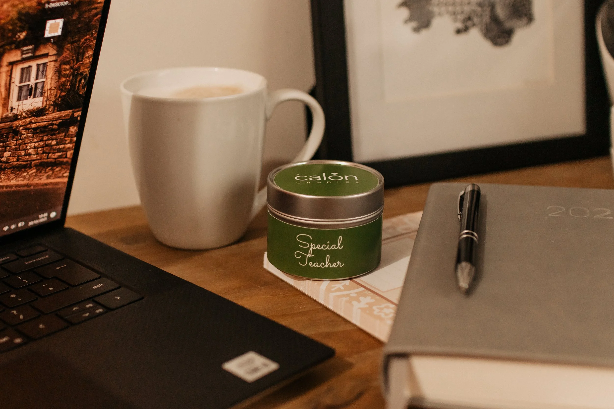 A desk with a laptop, a white mug filled with a hot beverage, a green mini tin candle labeled 'Calon Candles - Special Teacher,' a closed notebook with a pen on top, and a framed photo or art piece in the background.