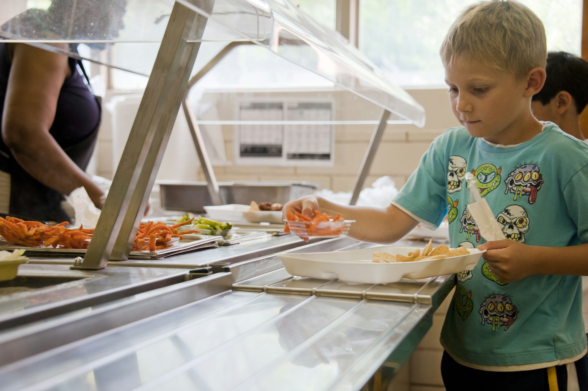 A young boy with blonde hair wearing a blue T-shirt with cartoon monsters is serving himself at a buffet with various vegetables, including carrots and celery, in a cafeteria-style setting.