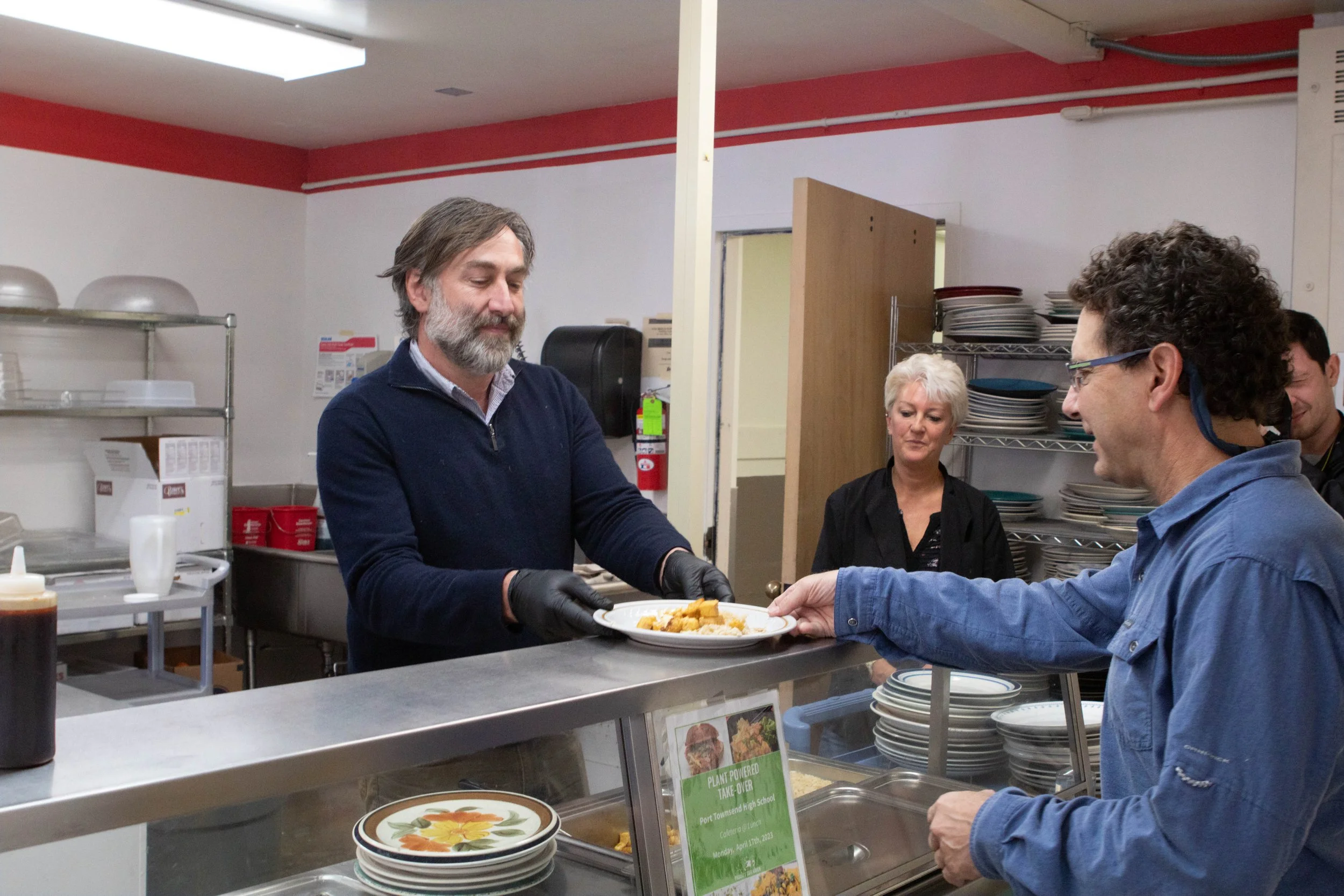 People in a cafeteria or kitchen serving food, with a man in a blue sweater and woman in black. The man is handing a plate of food to a man in glasses and a blue jacket. Shelves with dishes and a sign promoting plant-powered take-over are visible.