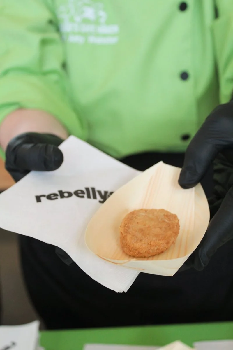 A person in a green uniform holding a piece of paper with the word 'rebellyous' and a paper tray with a breaded, fried food item on it, wearing black gloves.