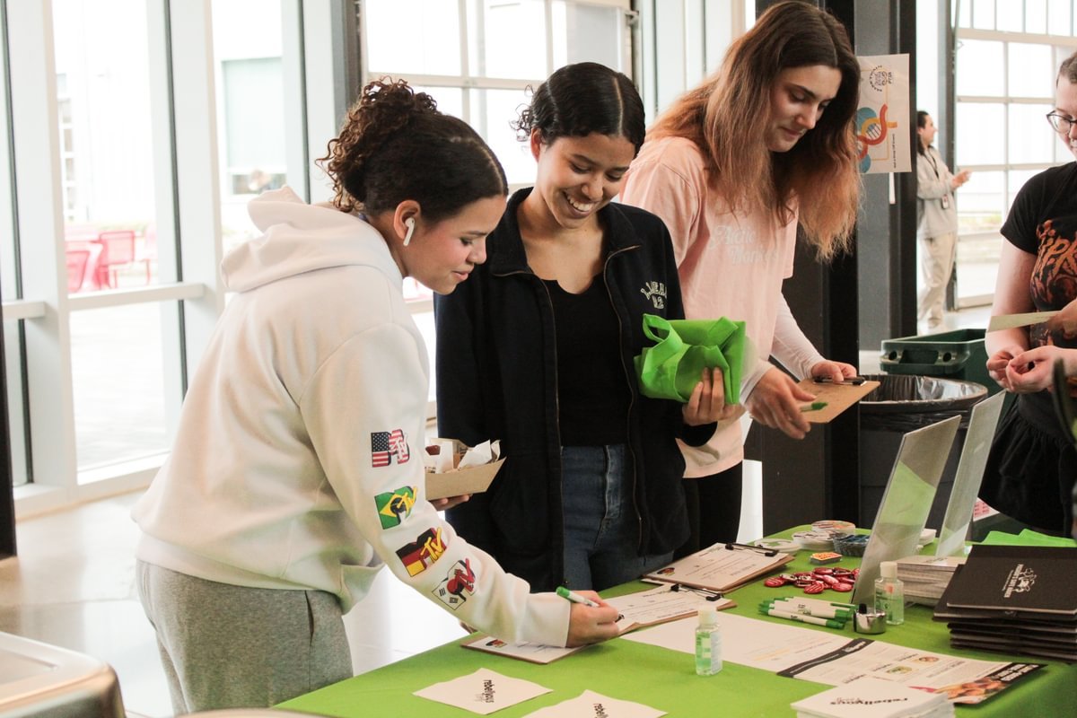 Group of women standing at a table inside a building with glass windows, engaging in a promotional or registration event. One woman is writing on a sign-up sheet, and others are holding green reusable bags and pamphlets.