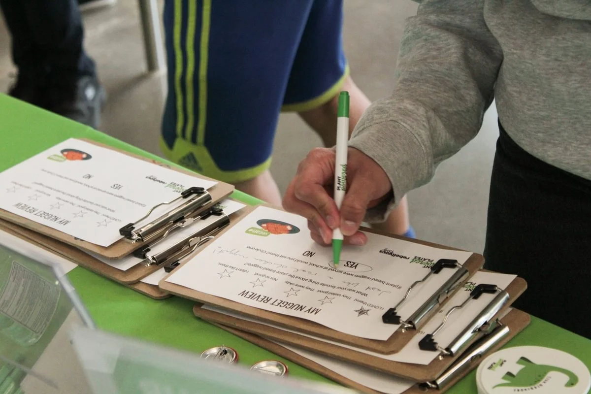 A person is signing a form on a clipboard at a green table, with additional clipboards nearby. Part of another person is visible in the background.