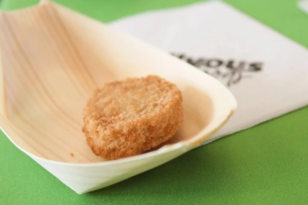 A breaded, round, fried food item placed in a white, boat-shaped dish on a green surface with a piece of paper underneath that has partially visible text.