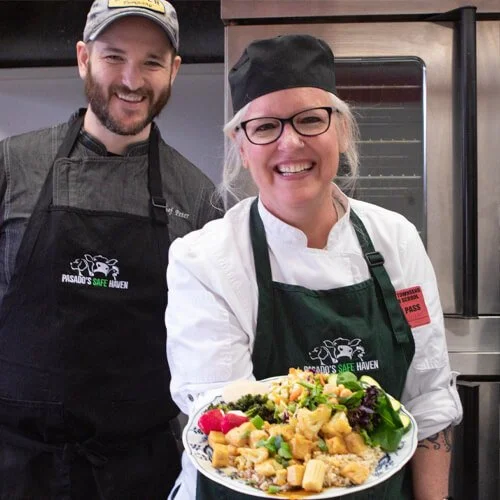 Two smiling chefs in black aprons and a woman in a white chef's coat holding a plate of colorful plant-based foods.