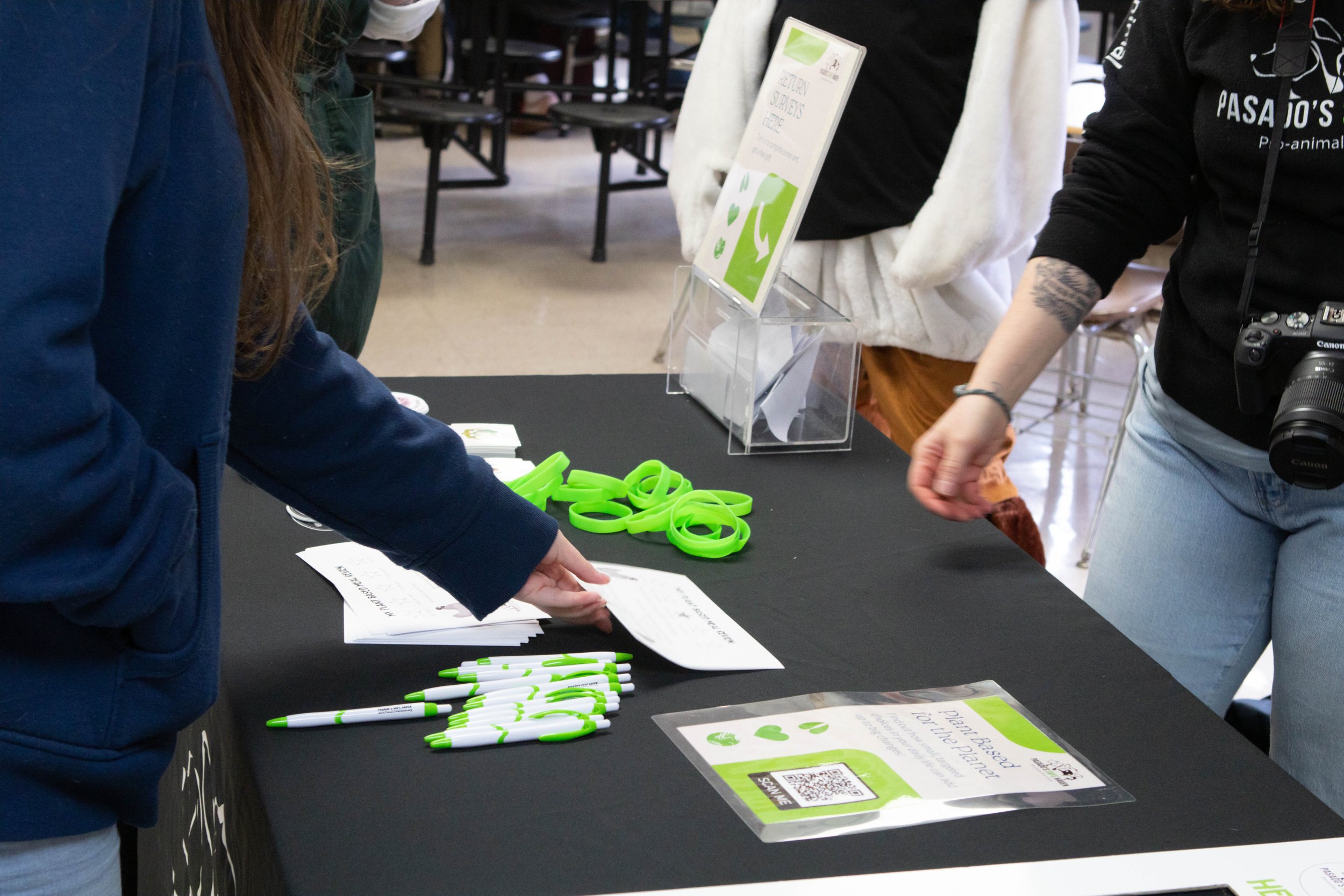A person at a table with green pens and papers, engaging with others at an informational booth about plant-based living, with a sign and informational materials displayed on the table.