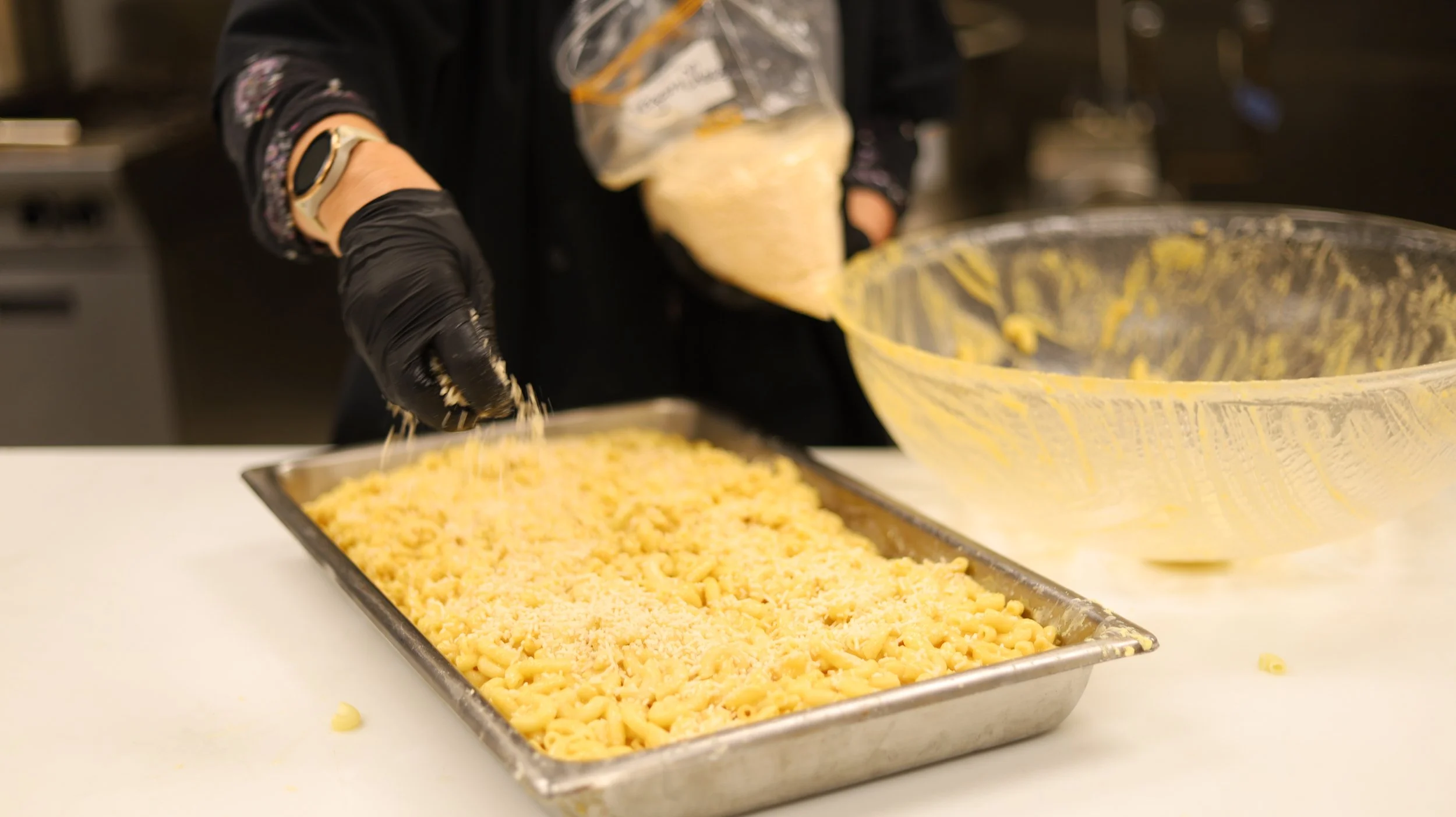 Person wearing black gloves and a black long-sleeve shirt is pouring shredded cheese from a plastic bag into a large glass mixing bowl. There is a metal tray filled with shredded cheese in the foreground. The scene takes place in a kitchen.