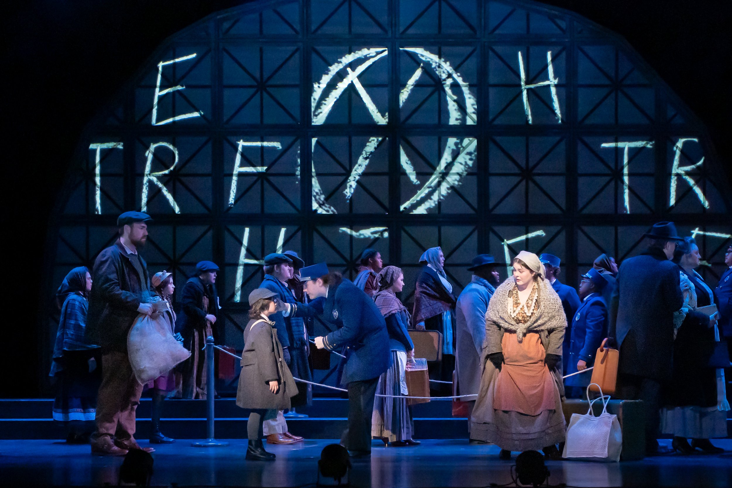 People dressed in period costumes in a "waiting room" as part of a performance with illuminated letters on a wall behind them.