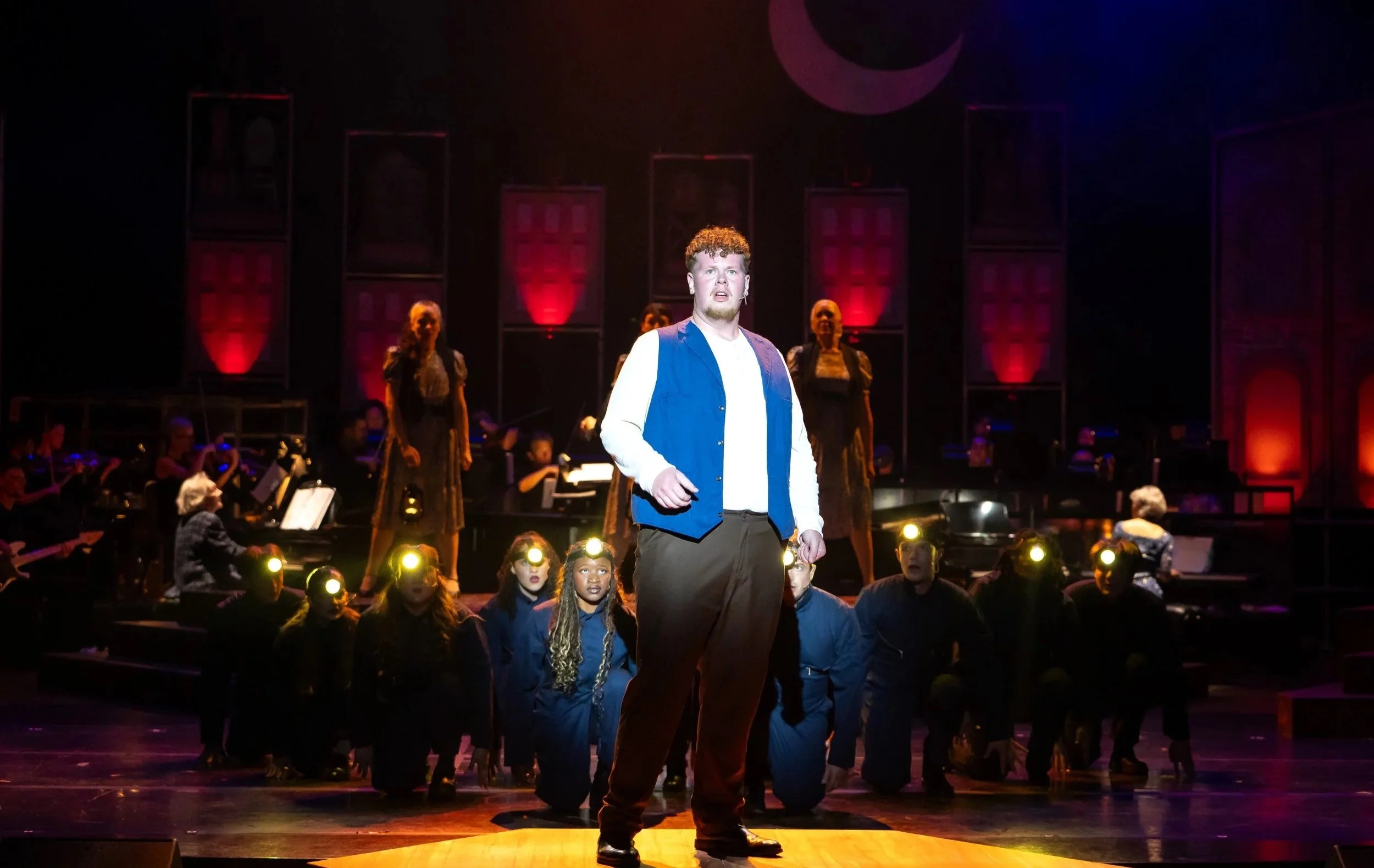 A stage scene with a man in a blue vest and white shirt at center, surrounded by others with headlamps and adults, with musicians behind him, and a dark backdrop with a purple crescent moon graphic.