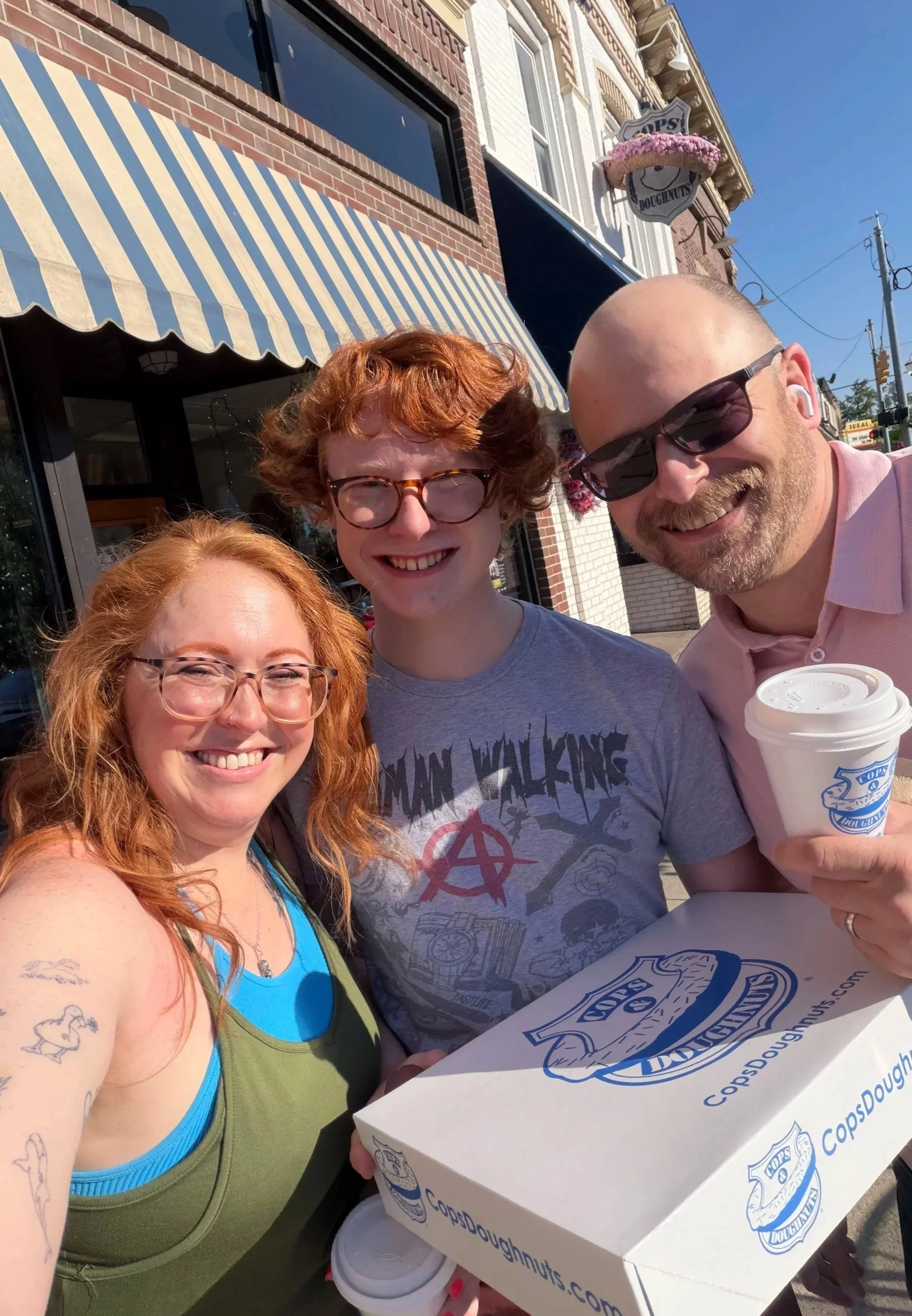 Three smiling people sitting outside a doughnut shop, holding a box of doughnuts and a coffee cup. One woman with red hair, a teenager with glasses and red hair, and a man with sunglasses, all enjoying a sunny day.