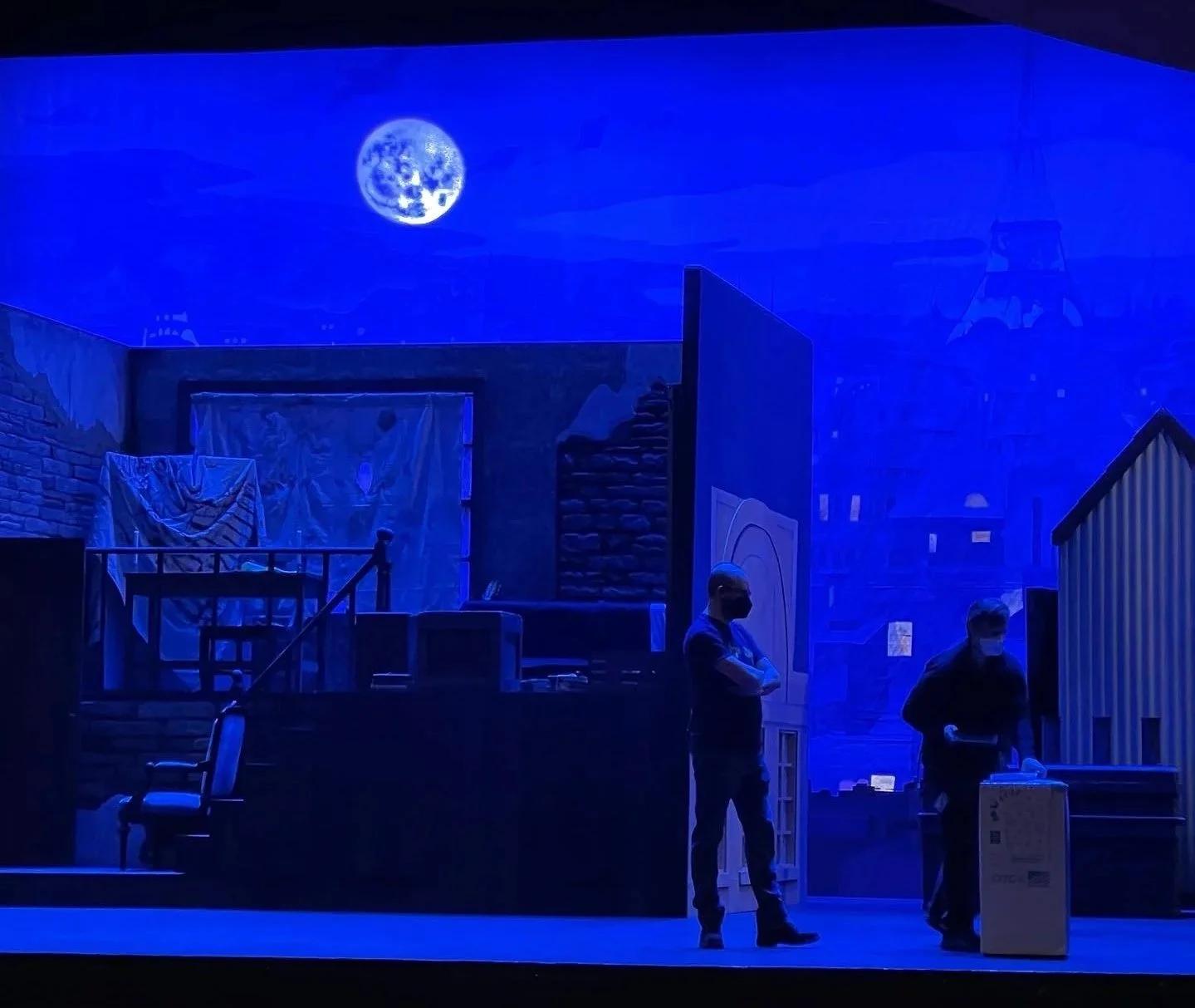 The stage set features a moonlit night sky with a full moon, a brick wall, a wooden railing with fabric draped over it, and two people wearing masks standing near a cardboard box, with some stage props and machinery around.
